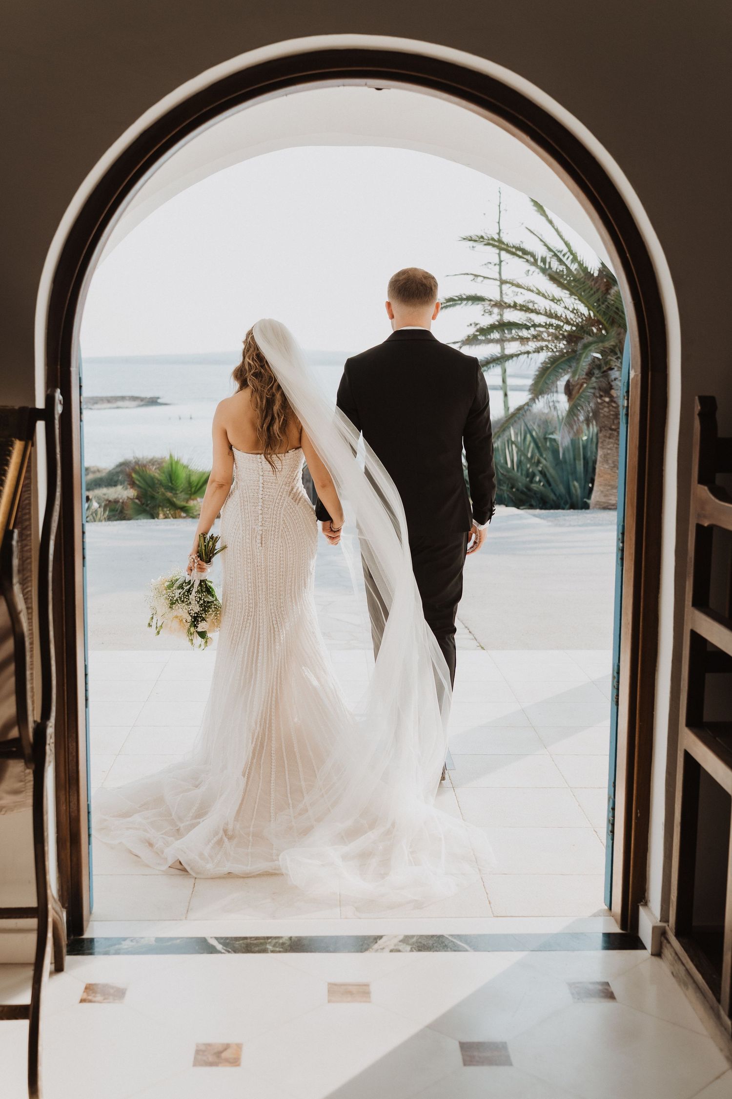 Bride and groom holding hands walking through arched doorway toward ocean view, bride in white gown with veil and bouquet.