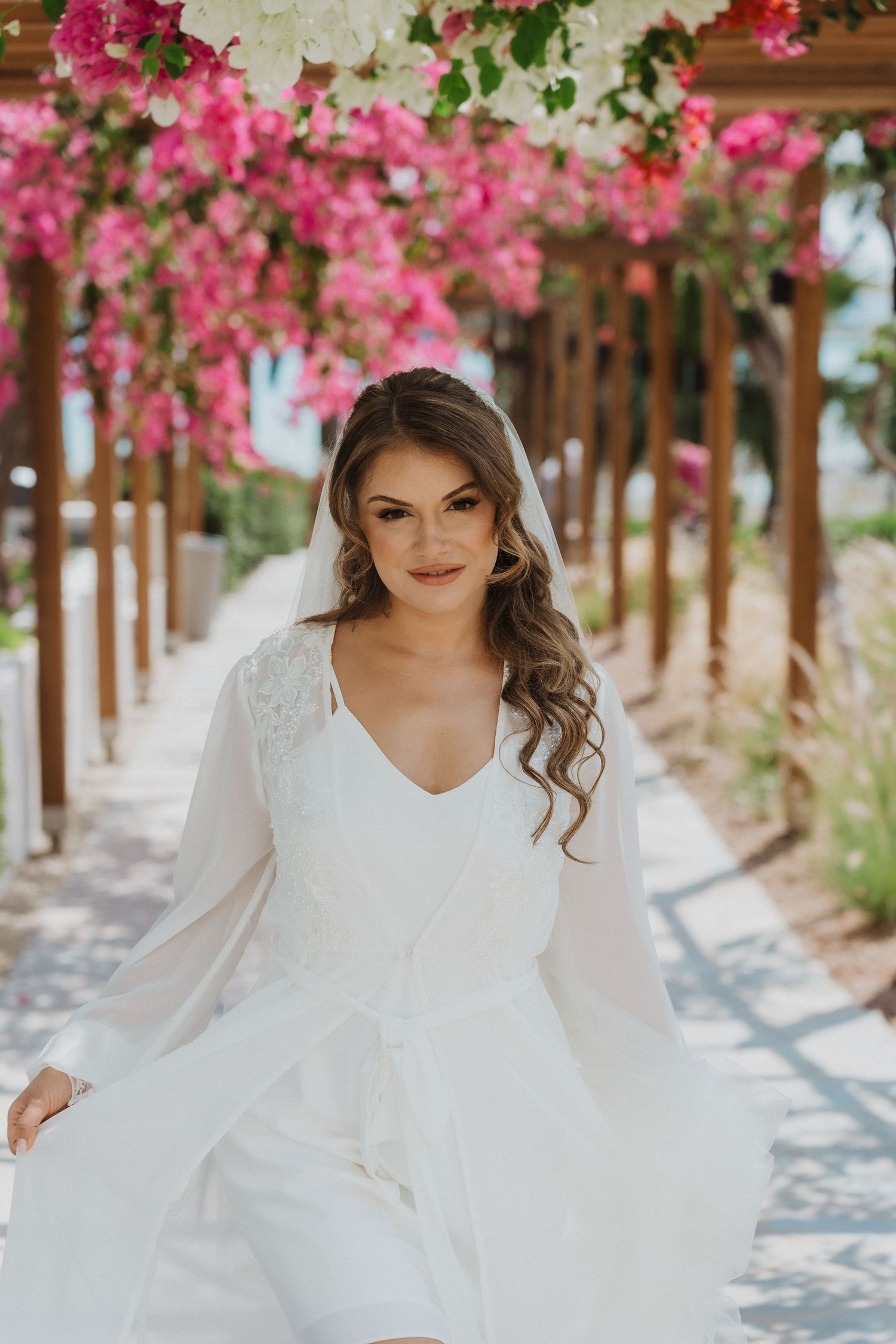 Elegant bride in white robe with veil posing under pink bougainvillea.