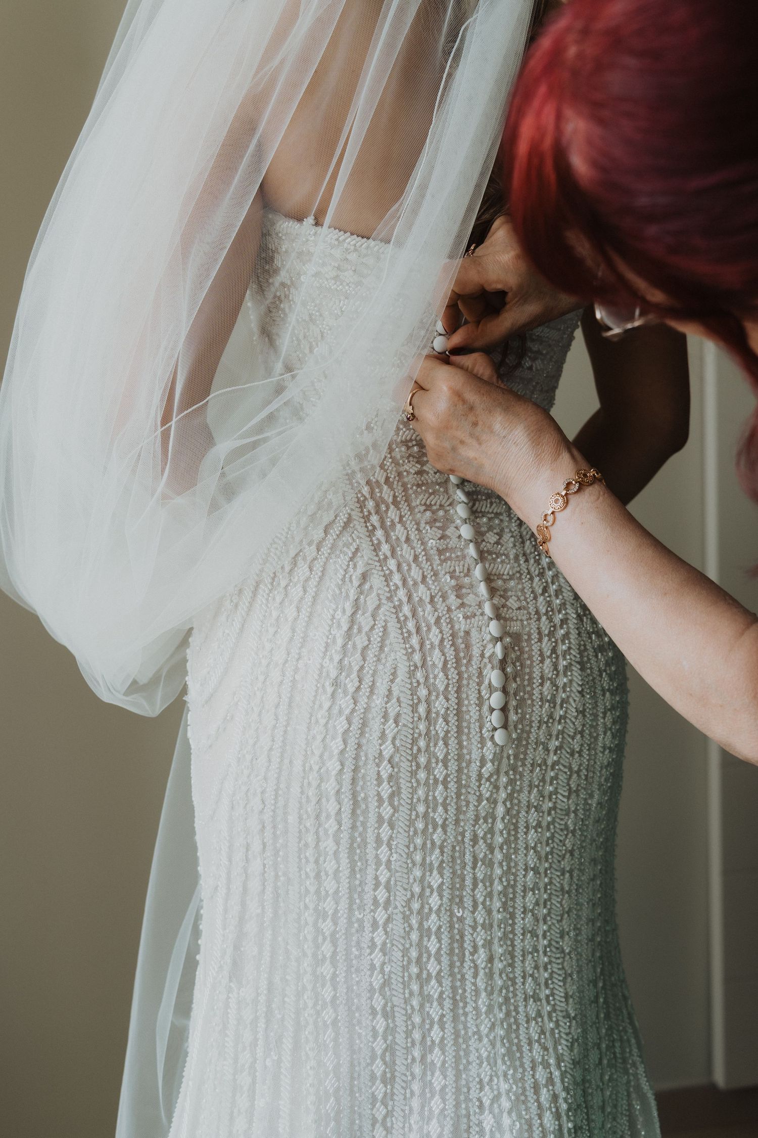 Bridesmaid with red hair fastens pearl buttons on brides fitted lace wedding gown with veil.
