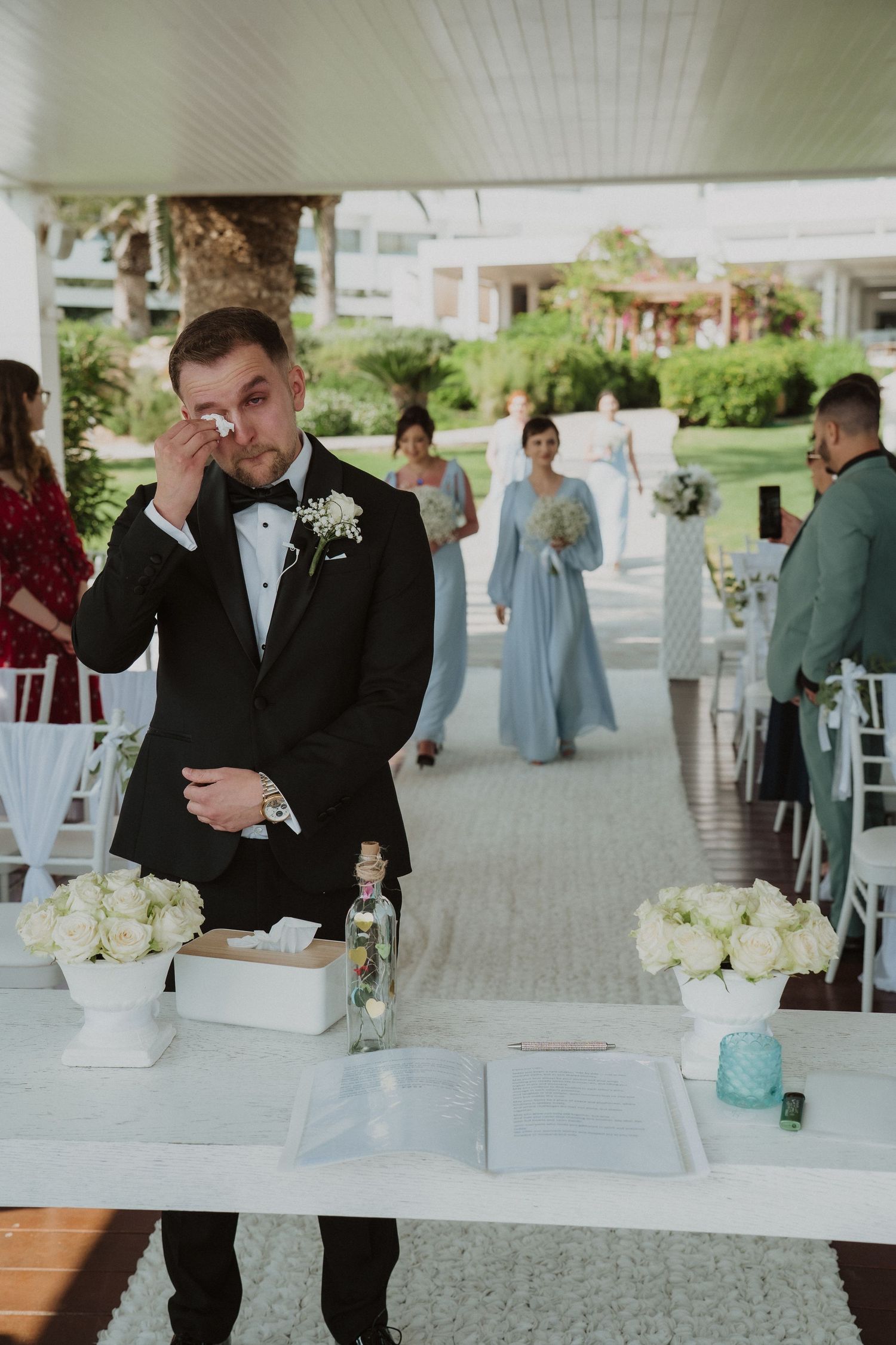Emotional groom in black tuxedo wiping tears as bridesmaids in blue dresses approach ceremony.
