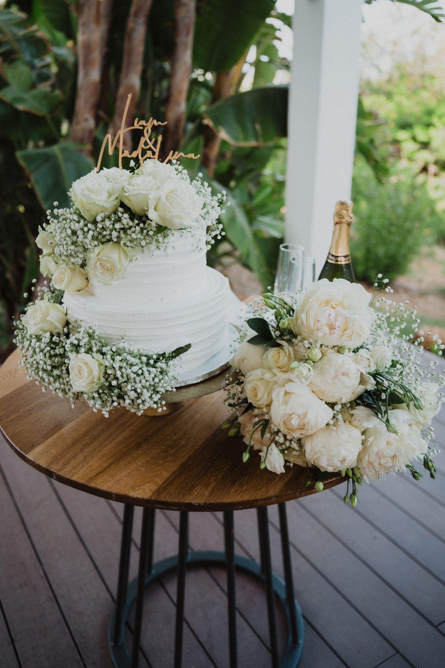 Two-tier white wedding cake adorned with roses and baby's breath beside white peony bouquet on wooden table.