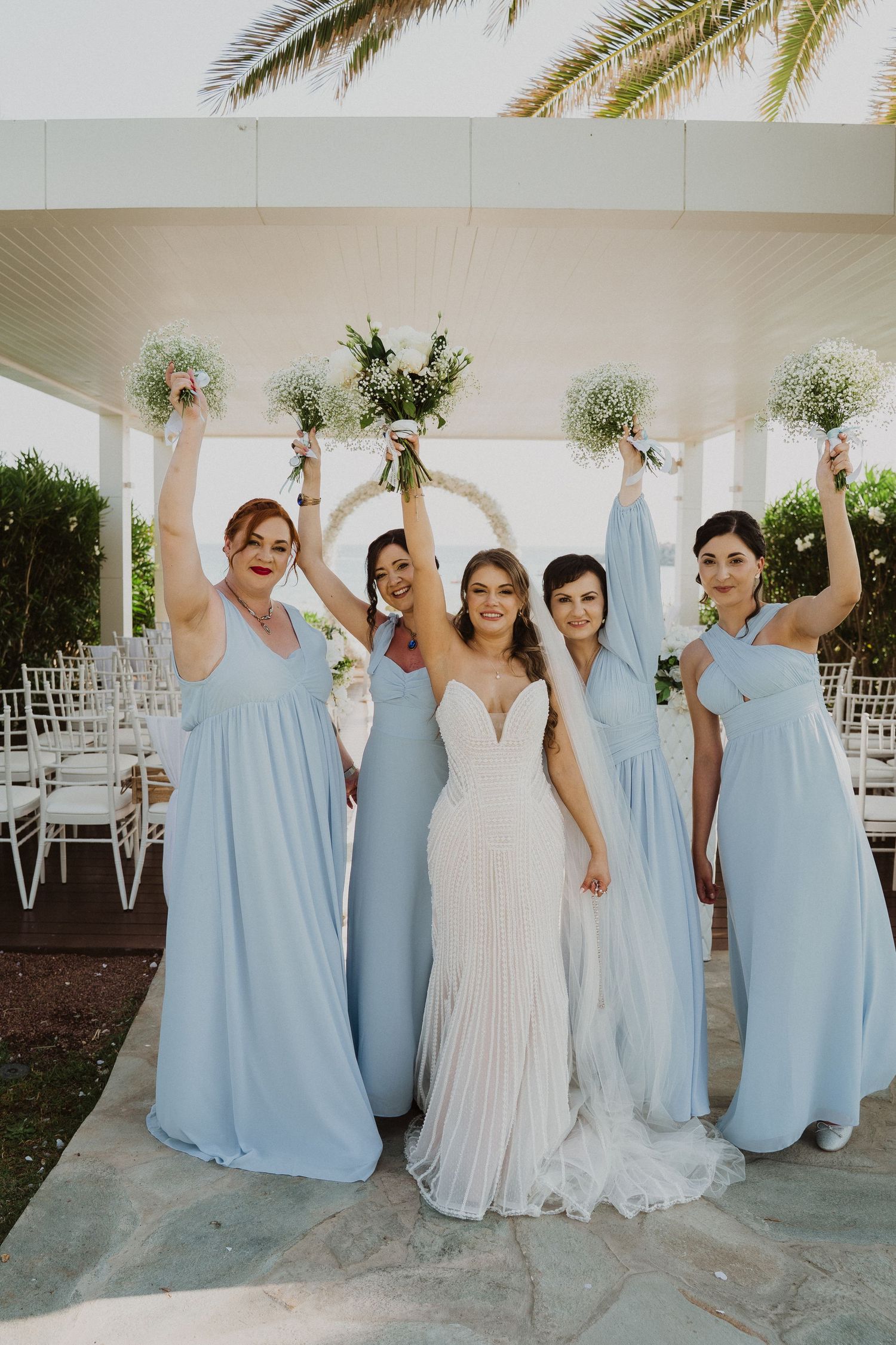 Bride in white gown with four bridesmaids in light blue dresses raising bouquets at outdoor venue.