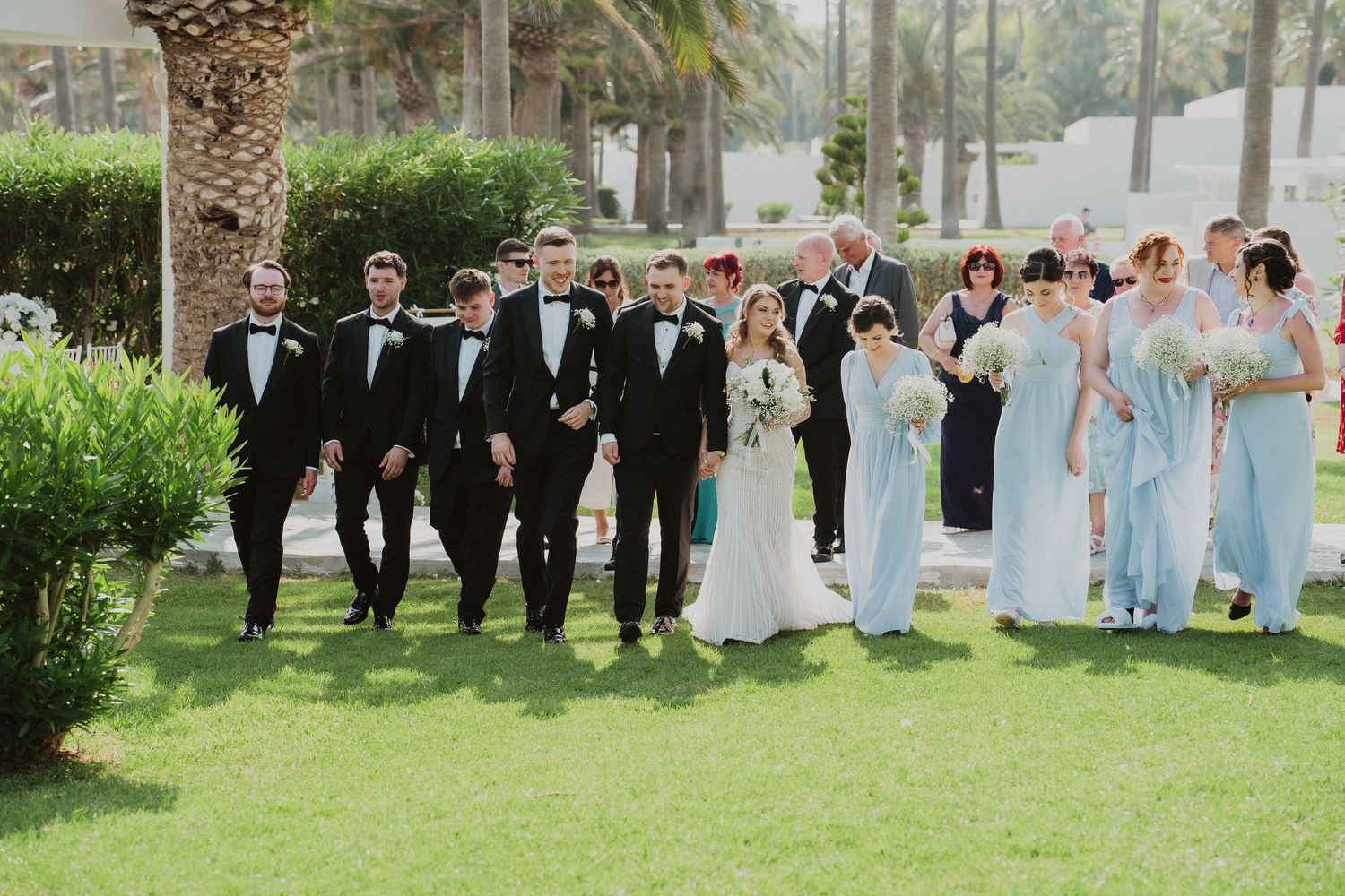Wedding party walks outdoors on green lawn, groomsmen in tuxedos and bridesmaids in light blue gowns with bride and groom.