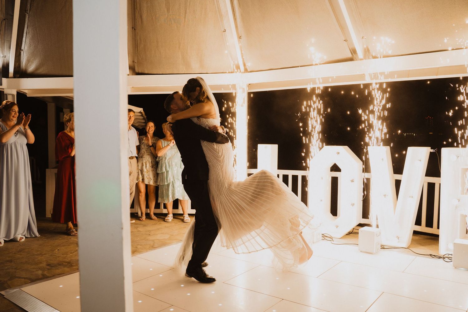 Groom lifts bride during first dance surrounded by sparklers with illuminated LOVE letters in background.