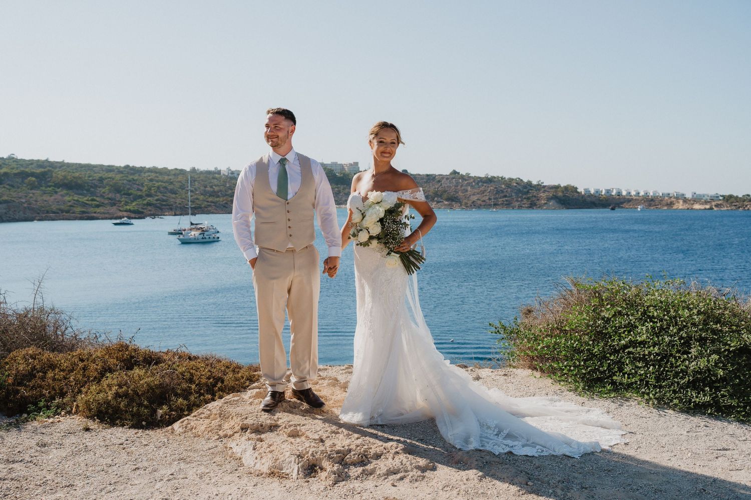 A wedding couple stands together overlooking a scenic coastal view with blue waters and rocky shoreline.