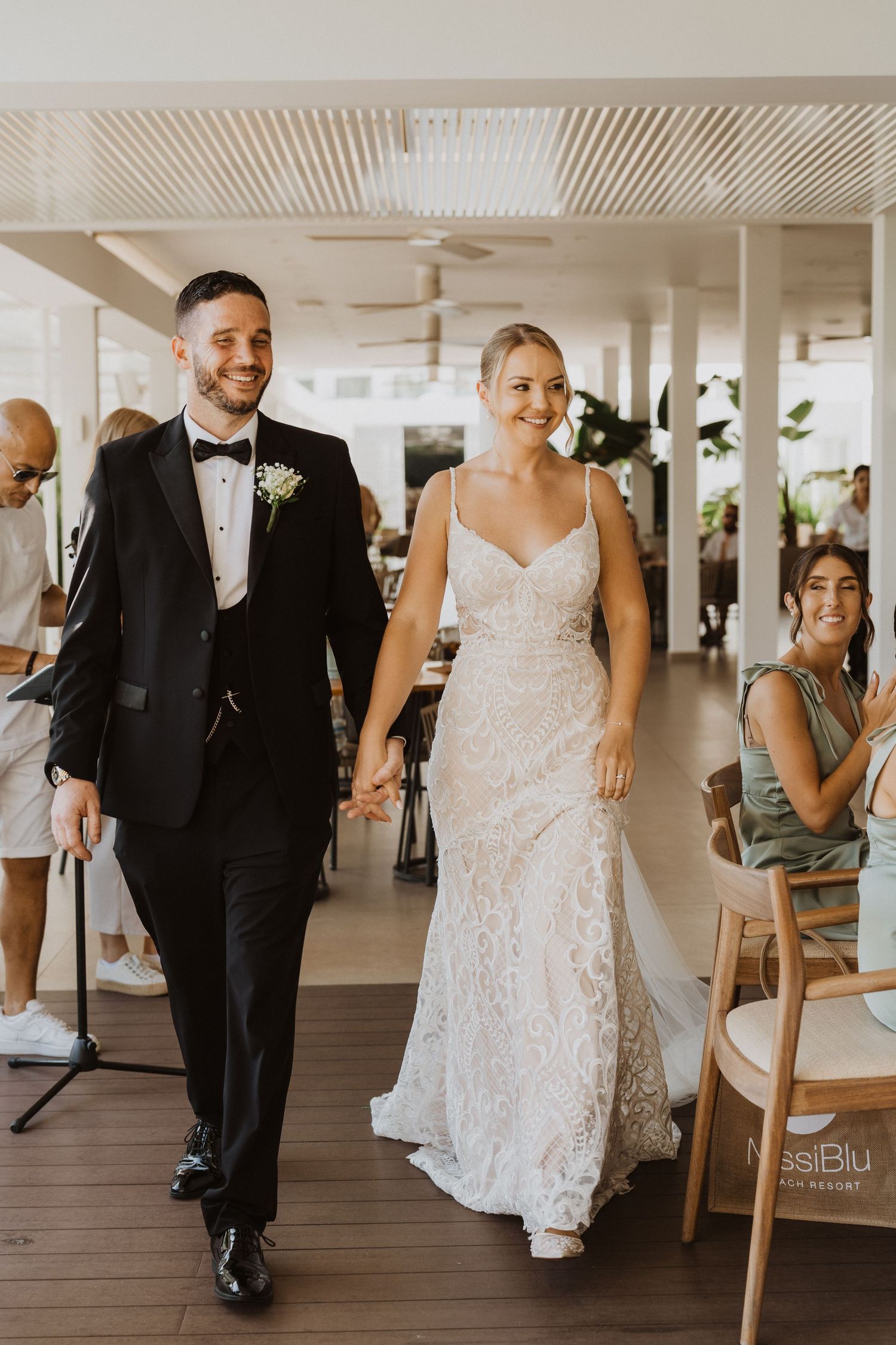 Wedding couple walks through an elegant indoor venue with white decor and wooden floors.