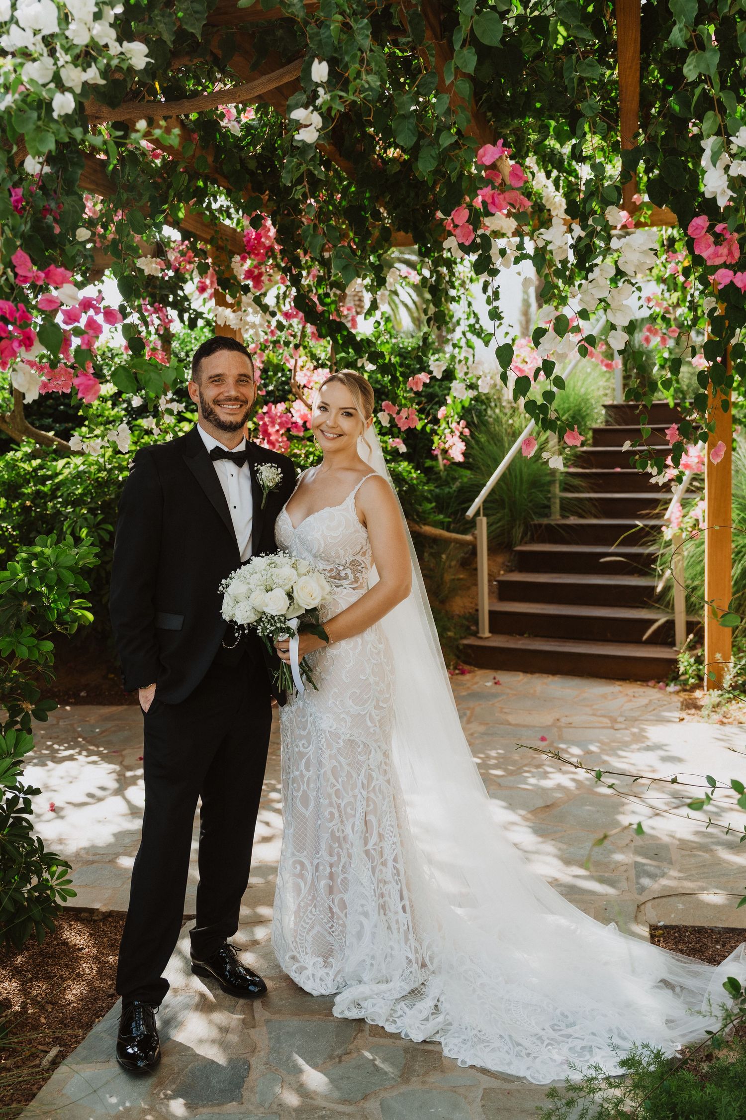 A wedding portrait captured on stone steps with lush floral archways of pink and white blooms overhead.