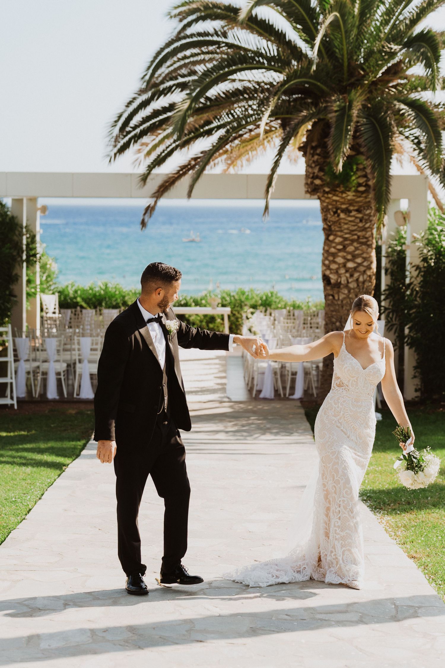A couple in wedding attire dancing together on a patio with palm trees and ocean views.