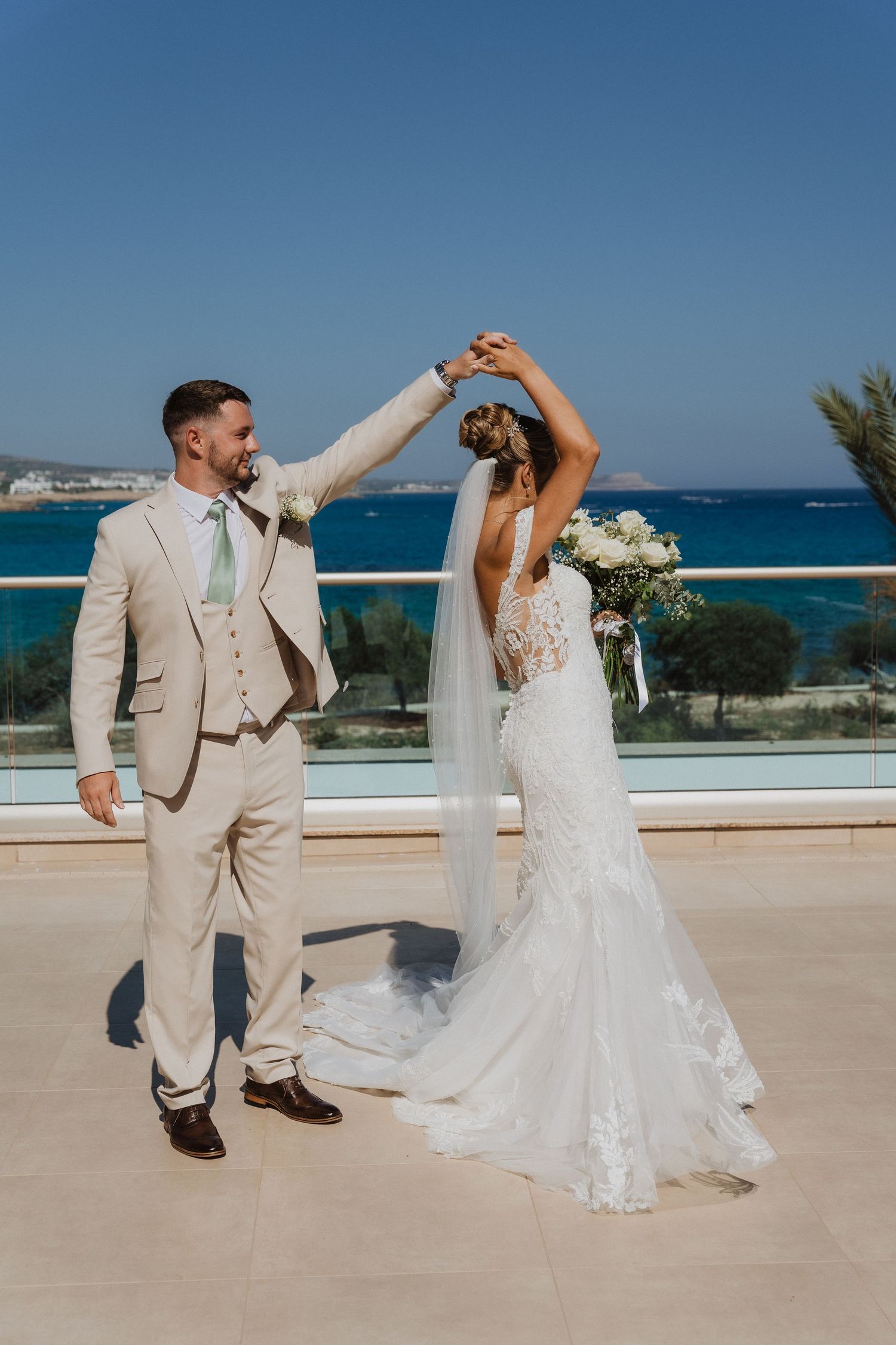A couple in wedding attire dances together on a beachfront terrace with ocean views in the background.