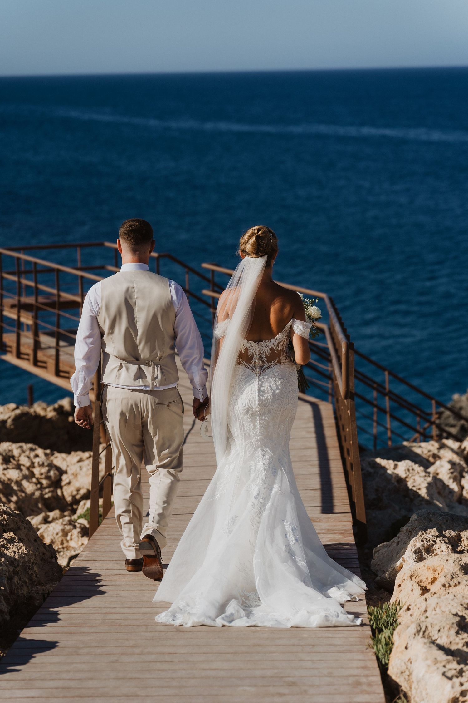 A bride and groom in white attire walk along a scenic coastal boardwalk overlooking blue ocean waters.