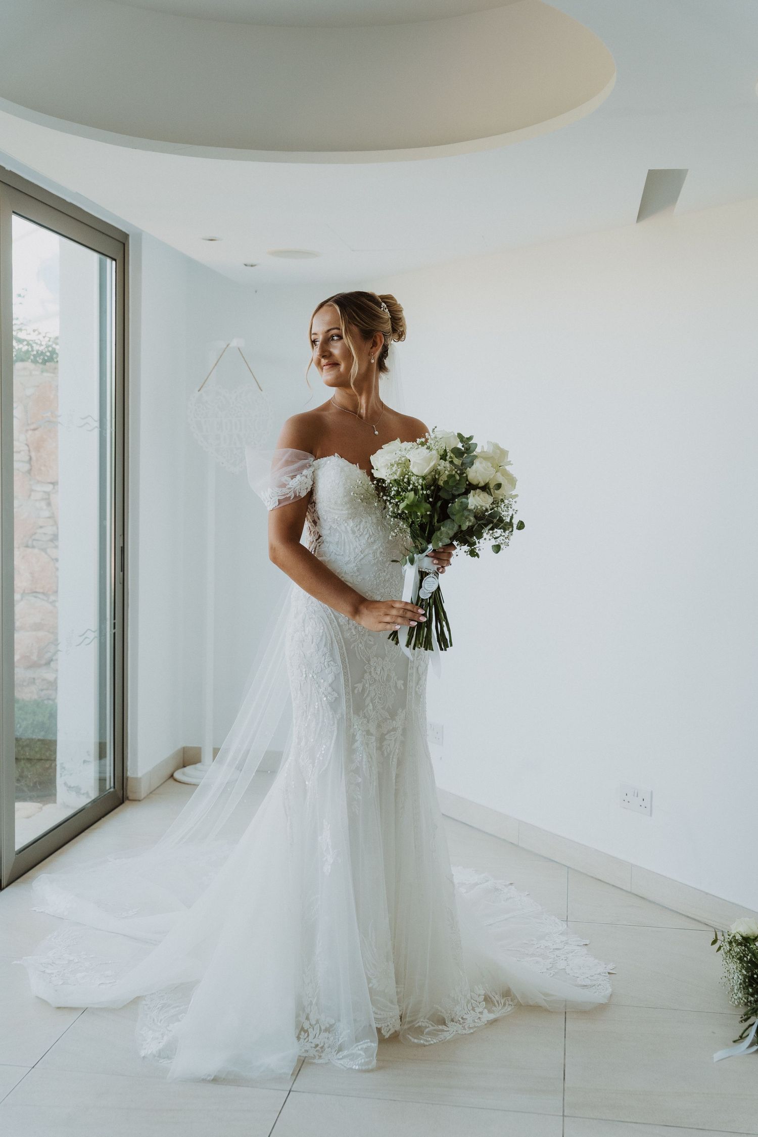 A bride in a flowing white off-shoulder wedding gown holds a bouquet of white flowers in a bright sunlit room.