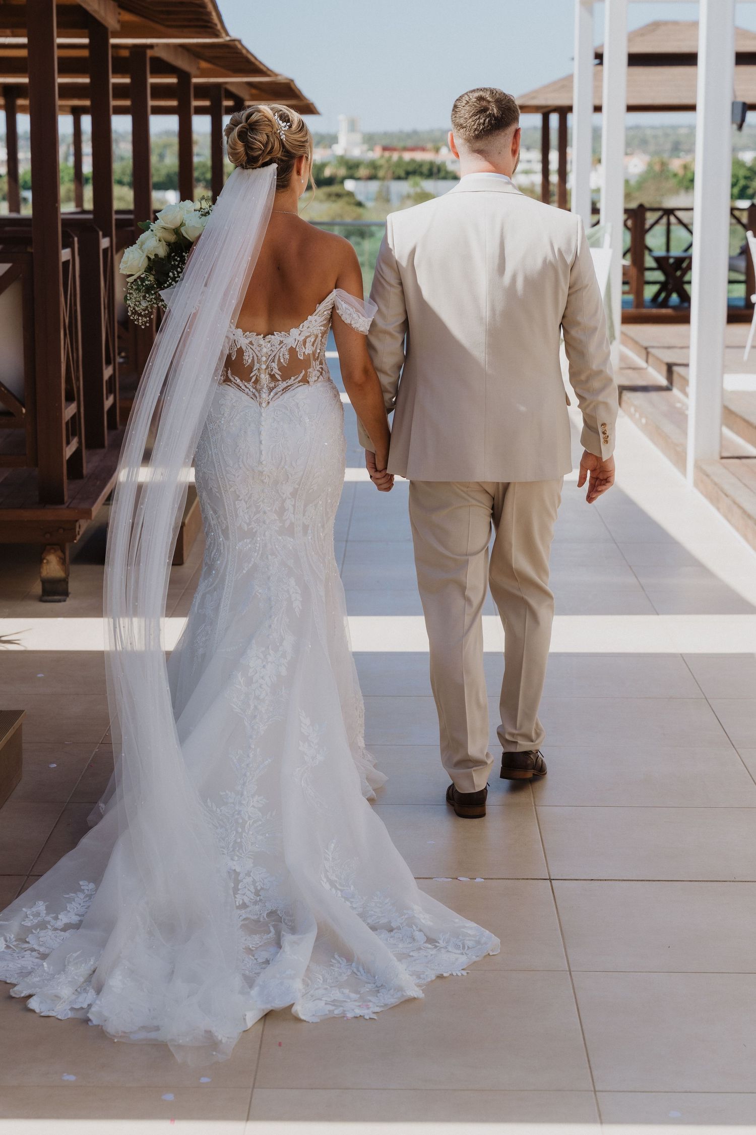 A couple in wedding attire walking on a covered outdoor terrace with white tile flooring and elegant decor.