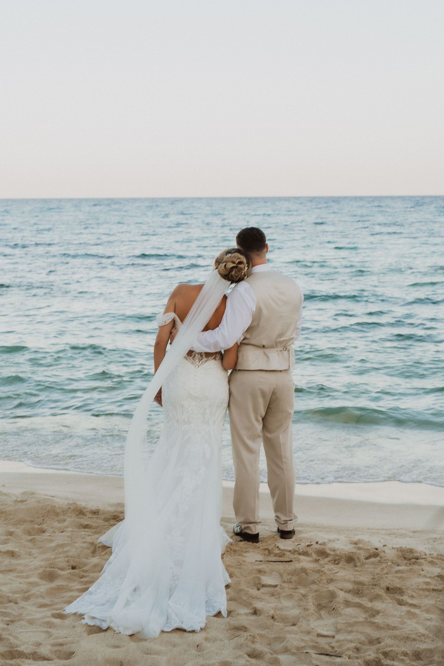 A couple in wedding attire embraces while standing on a sandy beach with turquoise ocean waves in the background.