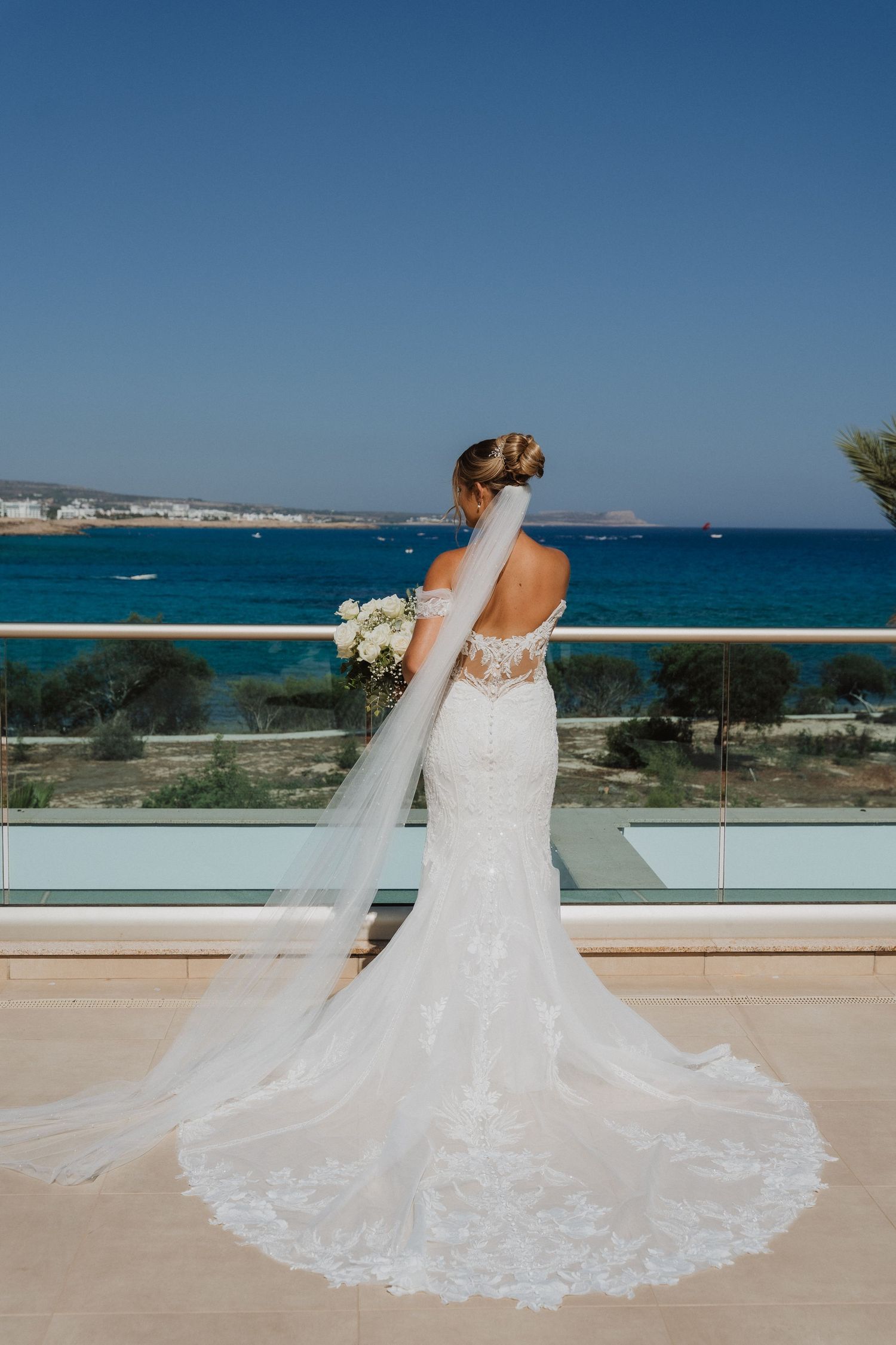 A bride in a lace wedding dress with a flowing veil stands on a balcony overlooking a stunning blue ocean view.