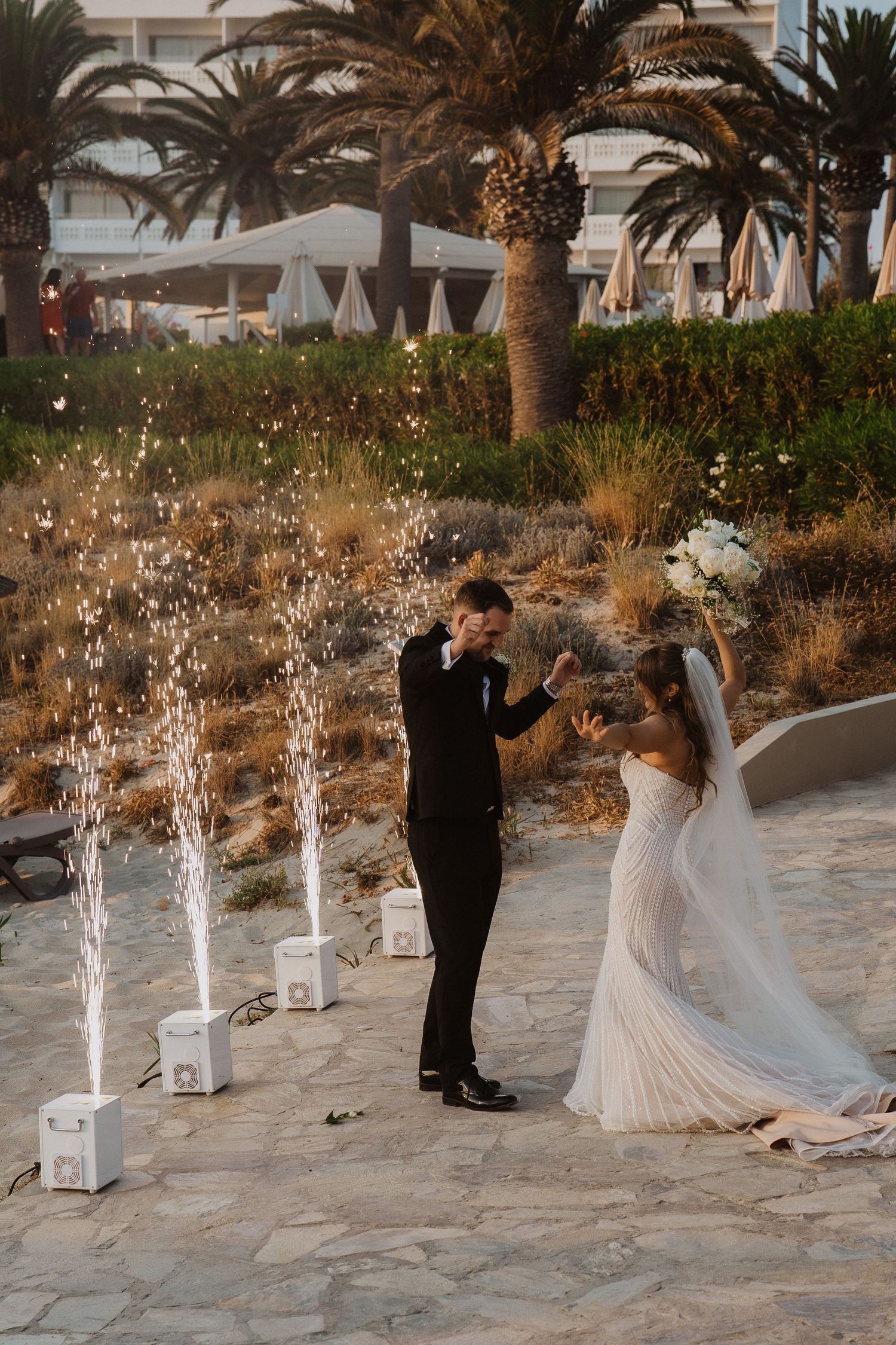 Bride and groom celebrate with cold sparkler fountains at outdoor wedding with palm trees at golden hour.