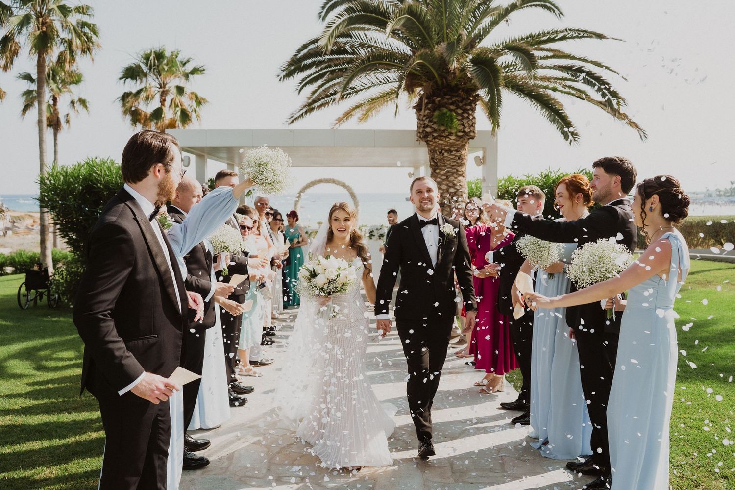 Bride and groom walk down aisle as guests throw confetti at outdoor wedding ceremony with palm trees.