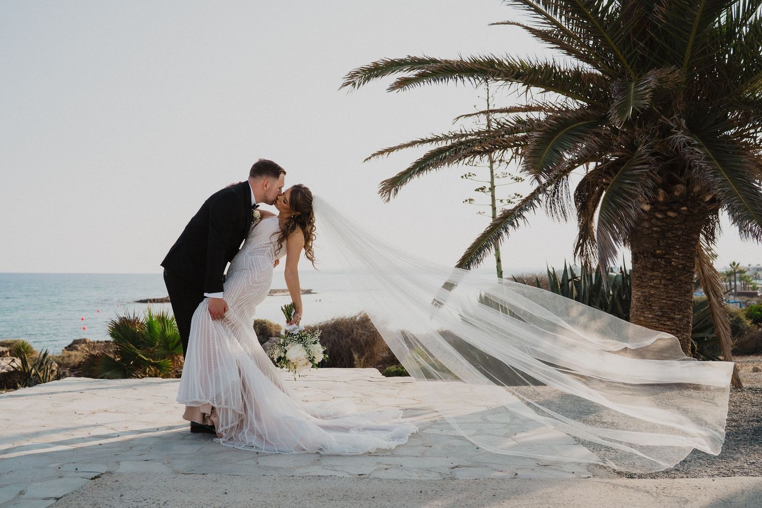 Groom dips bride for romantic kiss as her cathedral veil flows dramatically in ocean breeze.