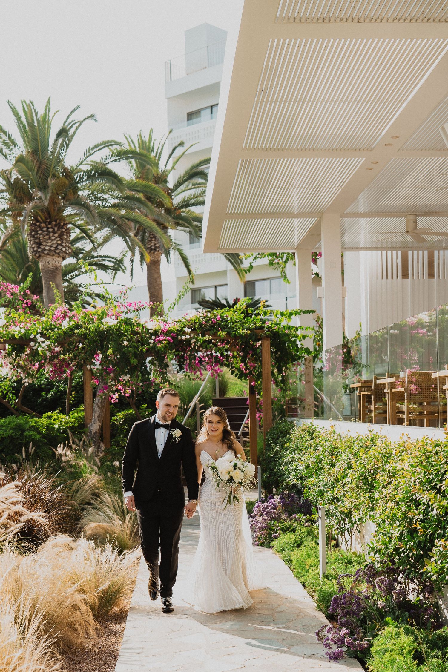 Elegant bride and groom walk hand-in-hand through lush tropical garden at luxury wedding venue.