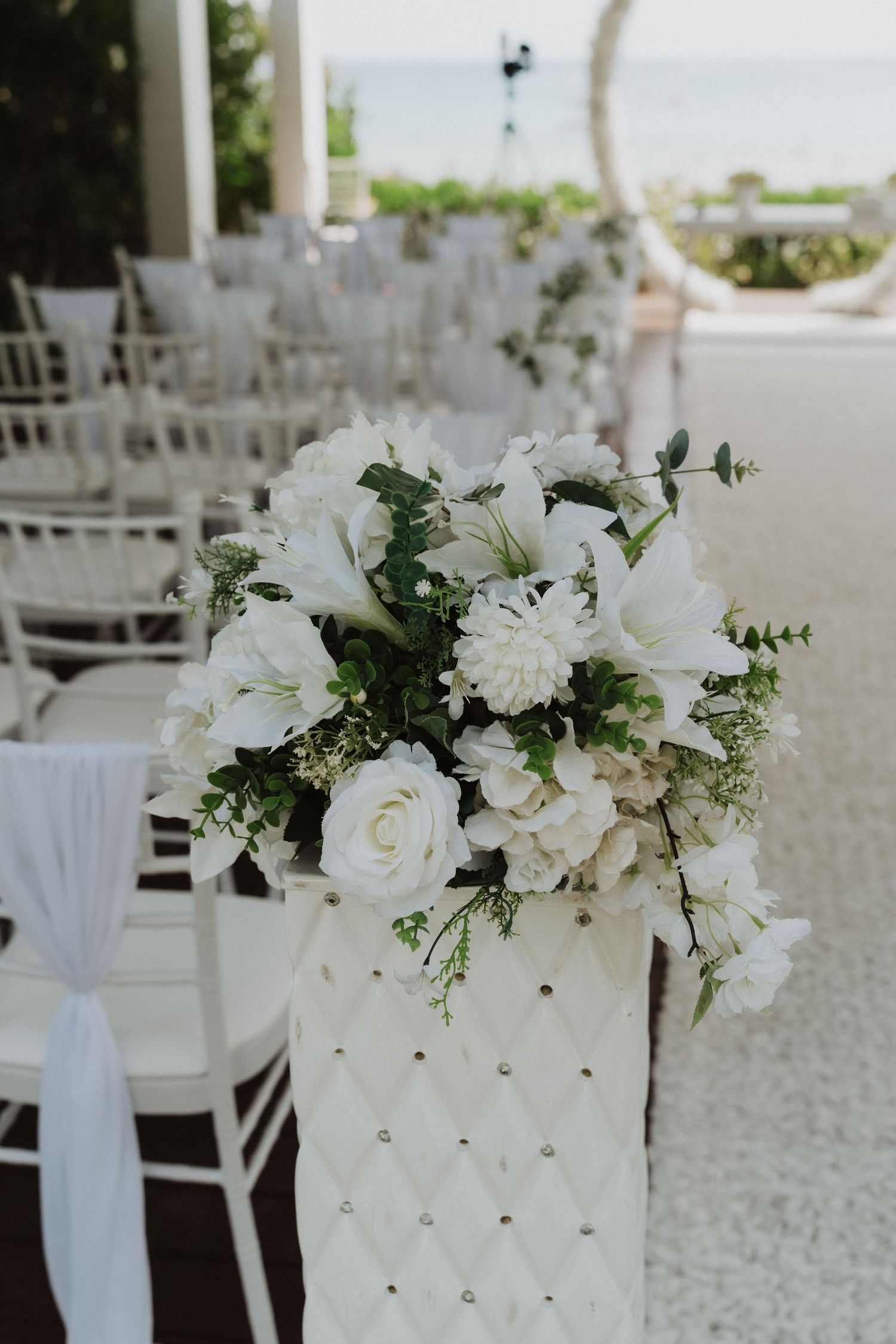 White floral arrangement with roses and lilies in quilted vase beside ceremony chairs near ocean.