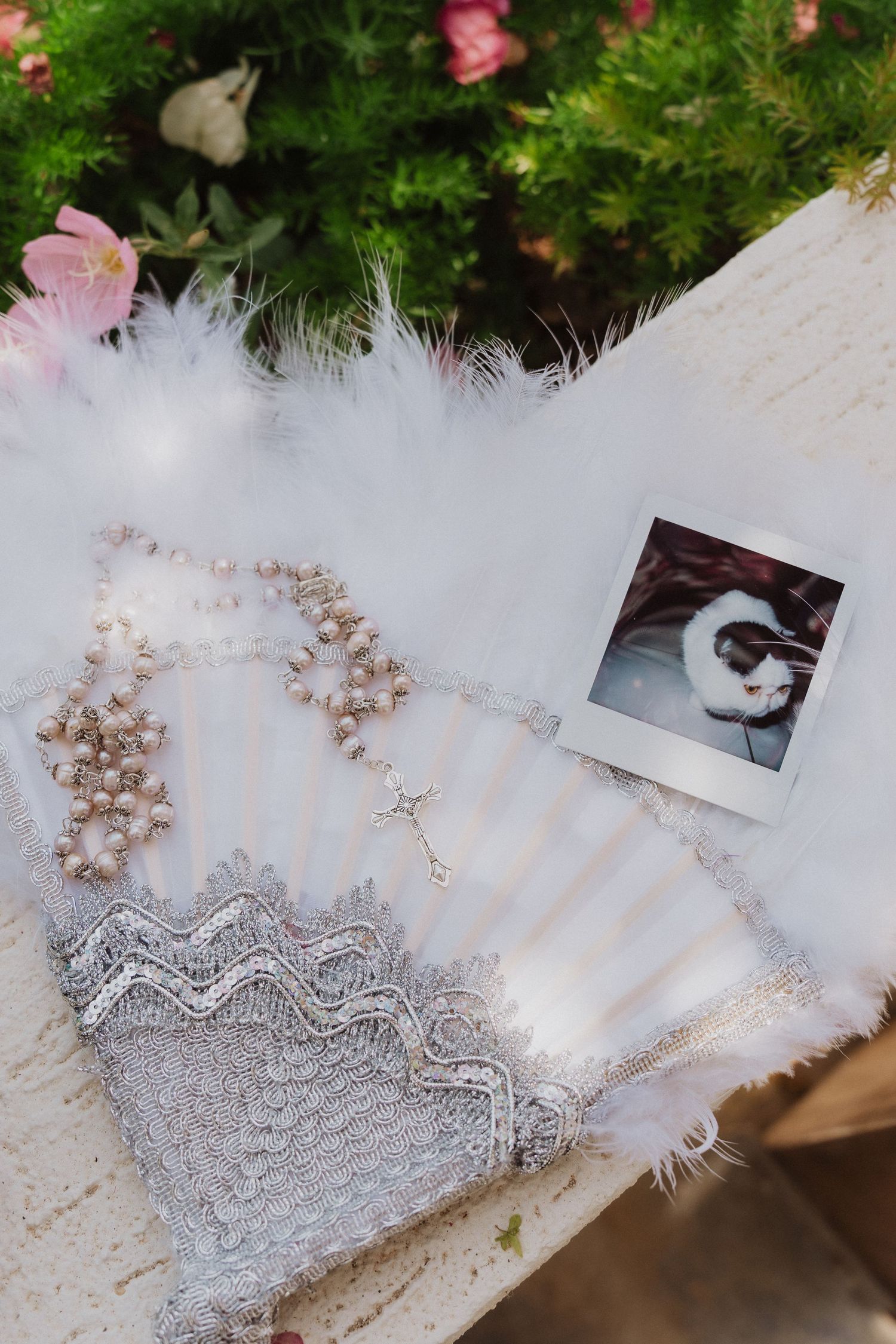 Bridal accessories flat lay with pearl rosary, lace fan, feathers, and Polaroid photo on white surface.