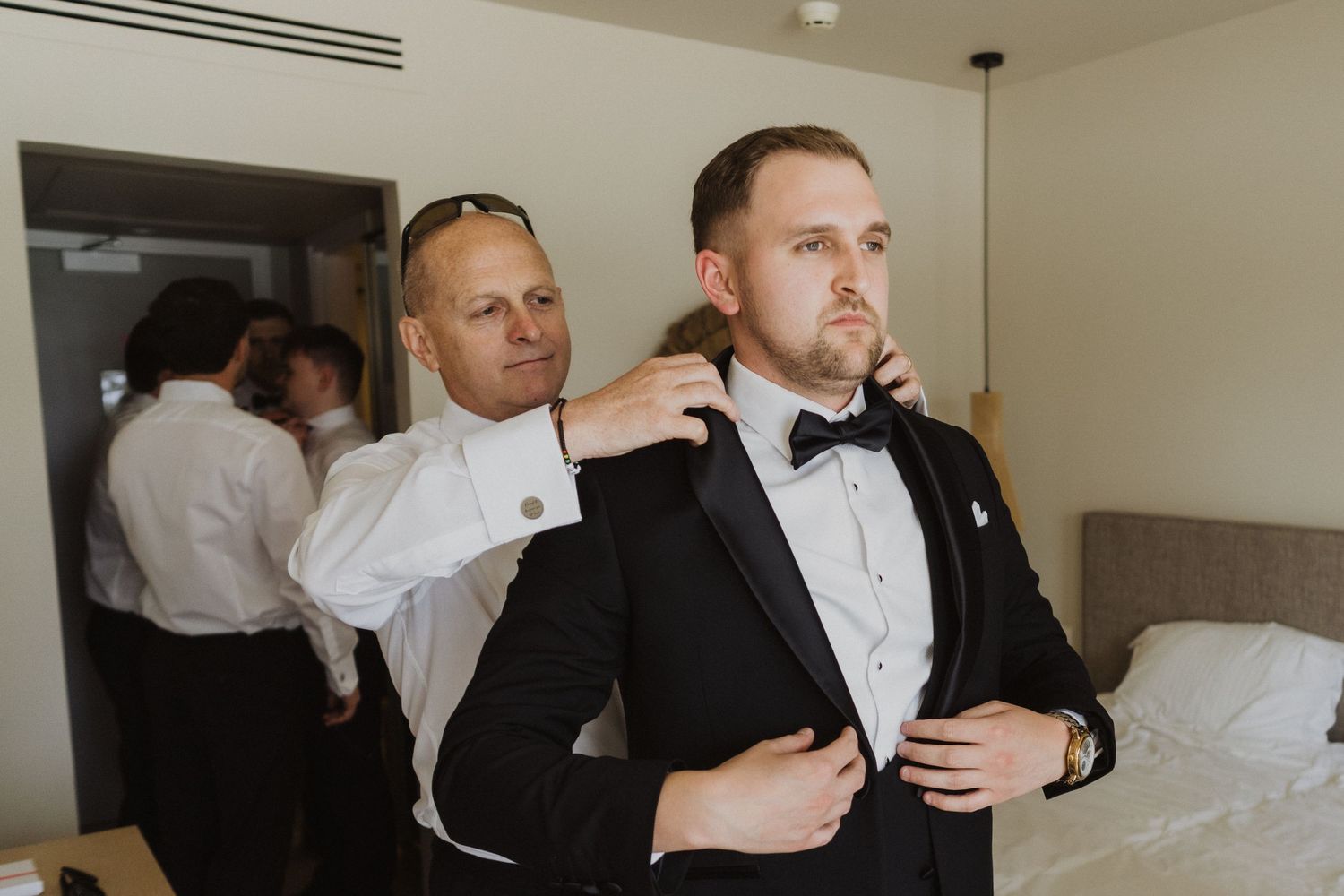 Groom in black tuxedo getting ready with groomsmen helping adjust jacket before wedding ceremony.