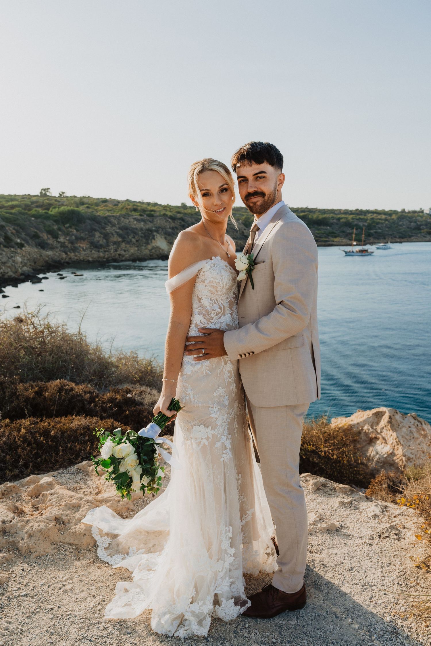 Seaside wedding portrait featuring elegant lace gown and light beige suit with coastal backdrop.