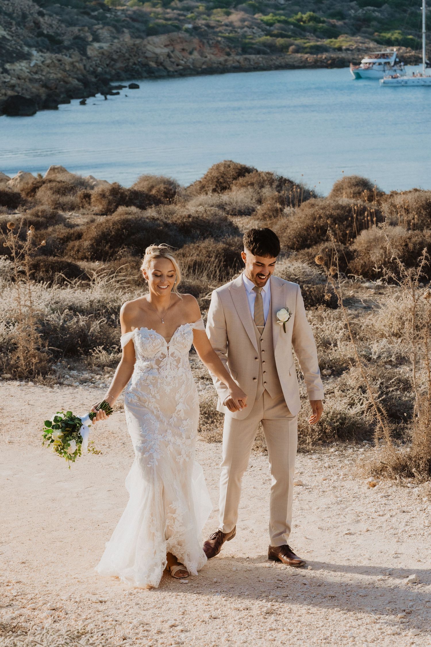 Bride and groom walk together on a sandy path with beautiful Mediterranean coastline in the background.
