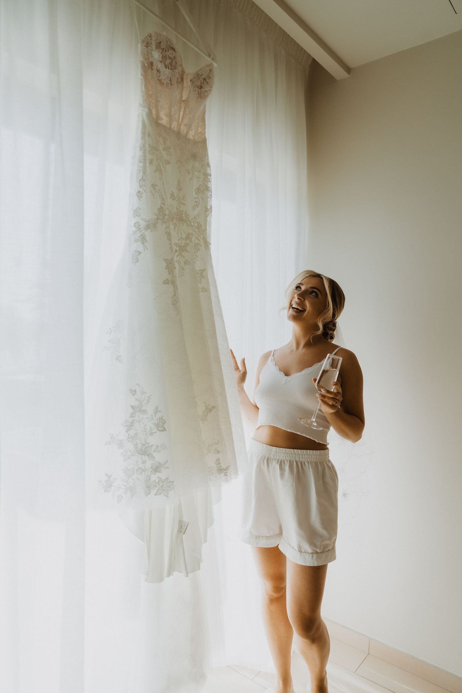 A delicate white lace wedding dress hangs by window while someone admires it in casual attire.