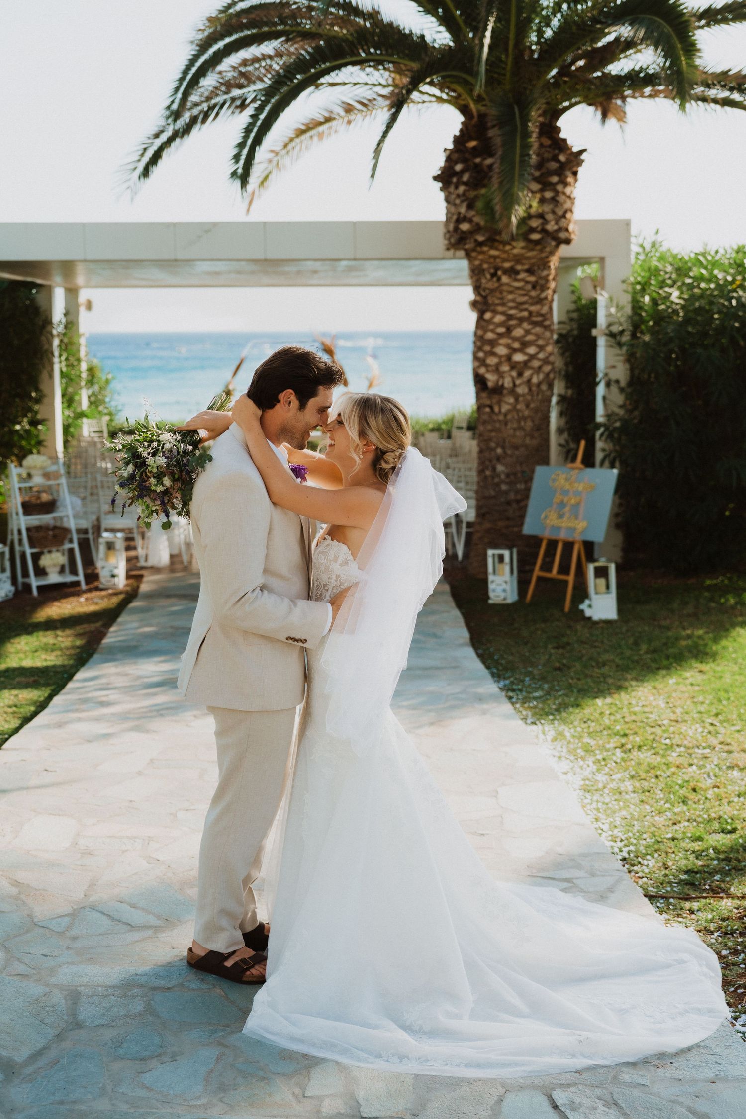Newlyweds dance together on stone patio with tropical palm trees and ocean in background.