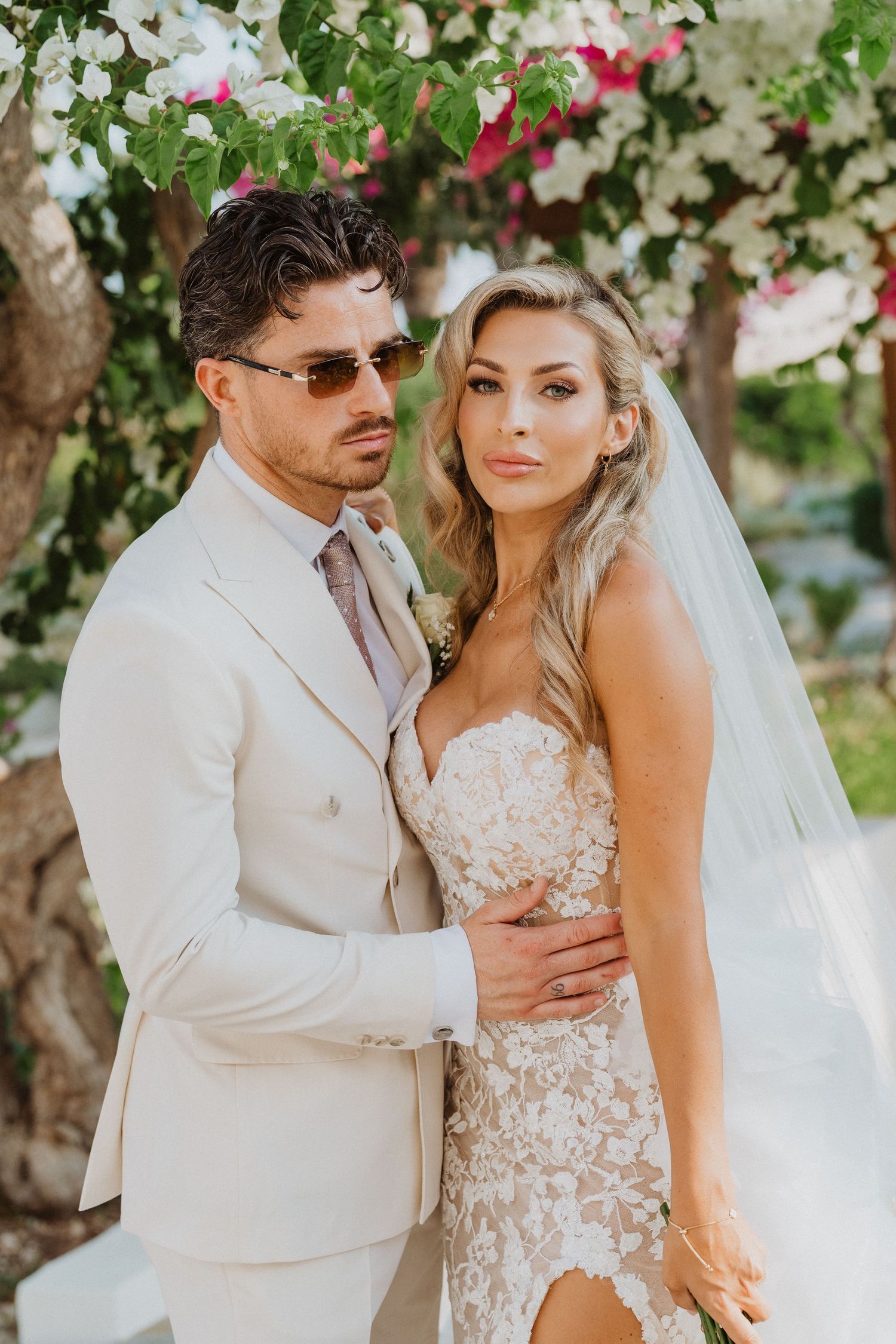A romantic wedding couple embraces outdoors among lush greenery wearing white formal attire and a flowing veil.