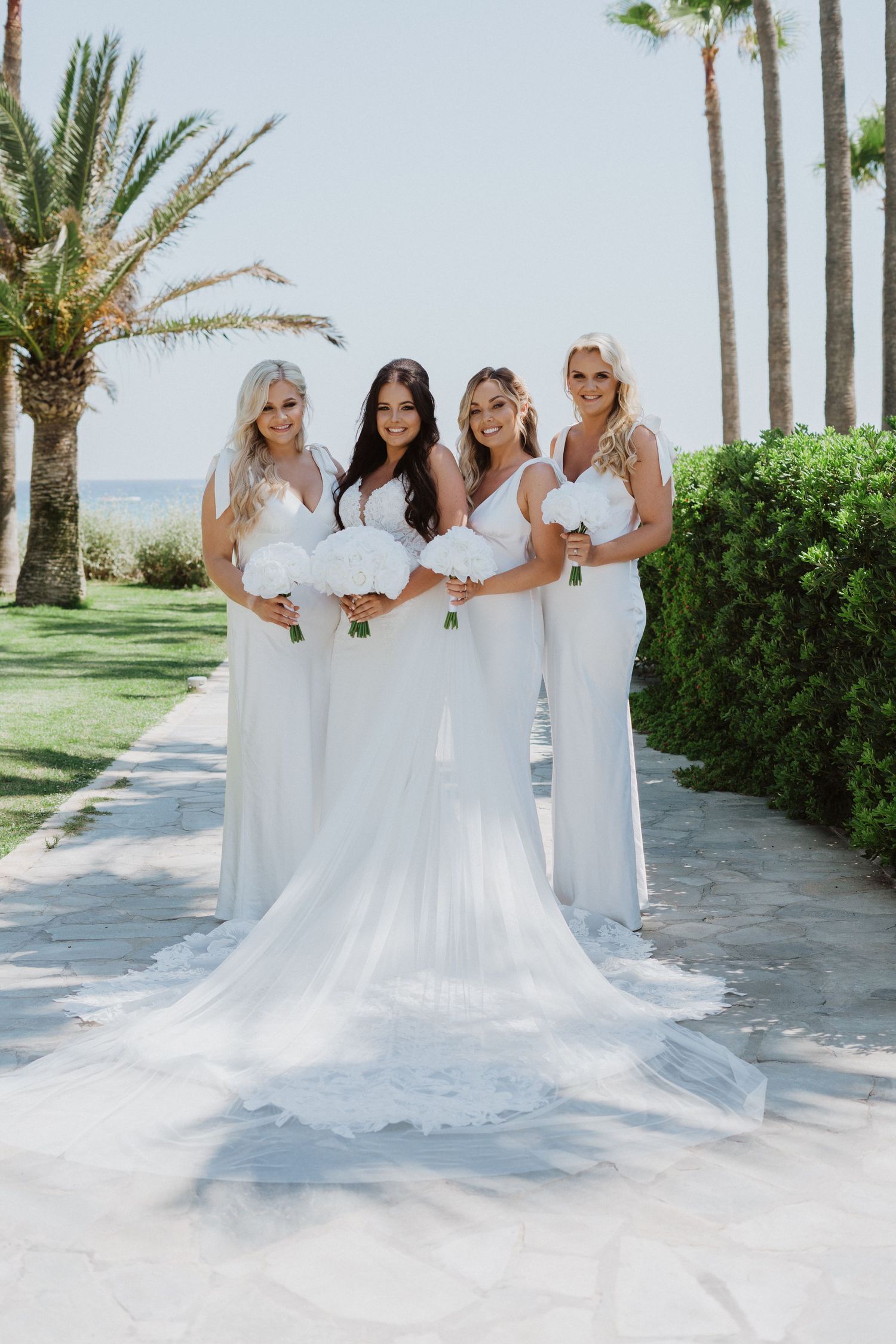 Bridal party in white dresses poses together on scenic oceanfront path.