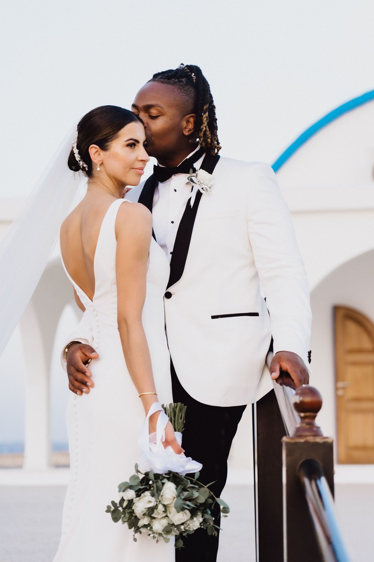 Romantic wedding portrait featuring elegant white tuxedo and flowing bridal gown against white church.