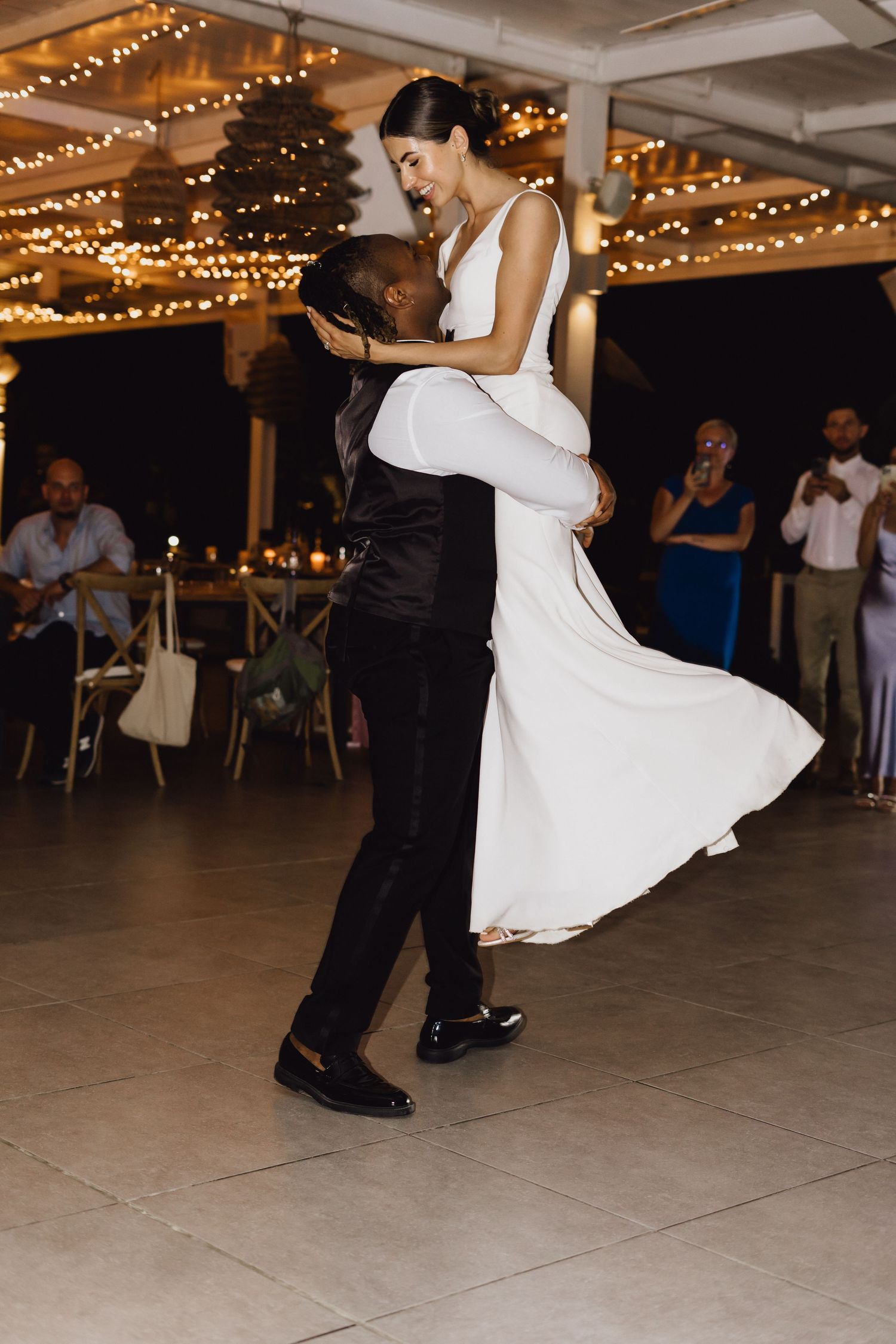 A romantic first dance lift at a wedding reception with string lights and guests watching in the background.
