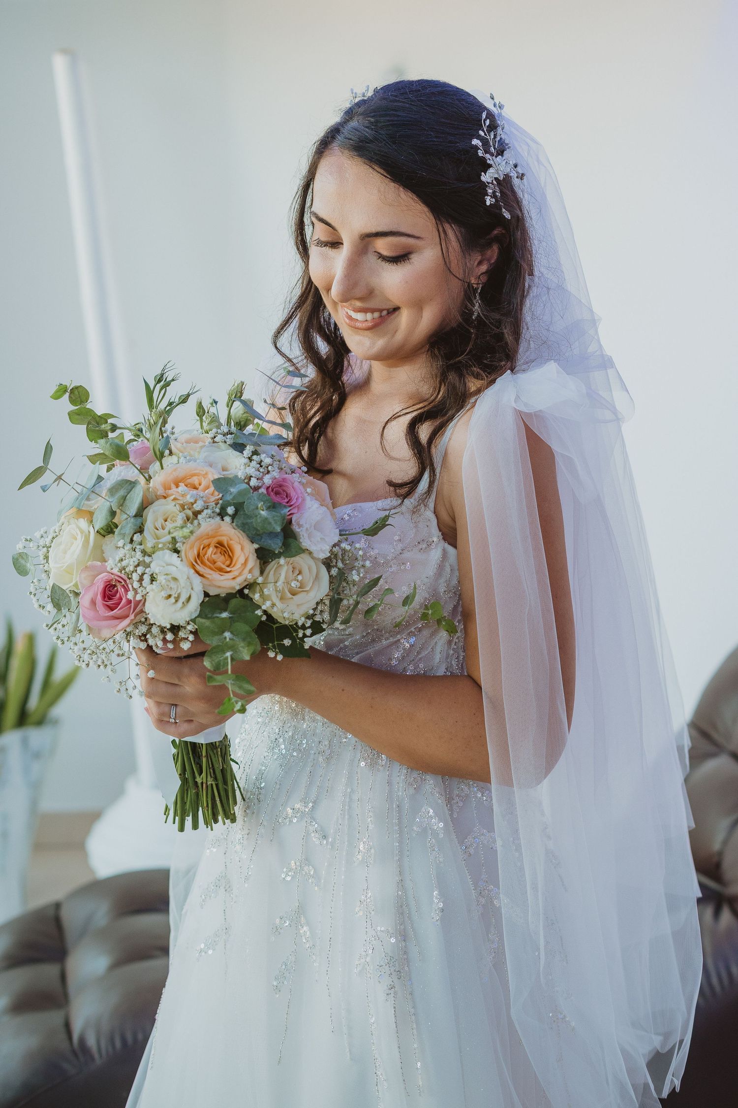 A bride in a white lace dress holds a beautiful pastel bouquet featuring roses and greenery while wearing a flowing veil.