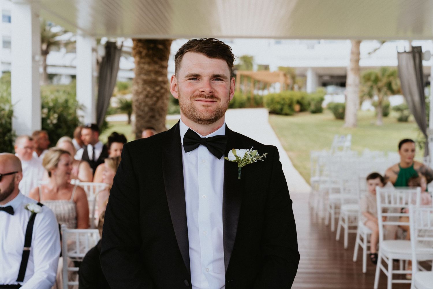Groom in classic black tuxedo standing at outdoor ceremony venue with guests seated behind.