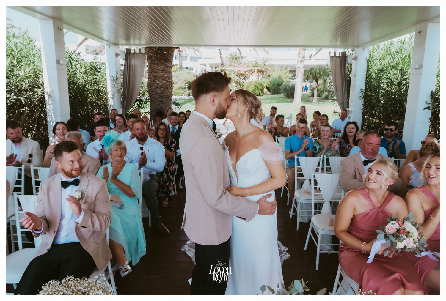 Celebratory kiss at wedding ceremony while guests watch from white chairs.