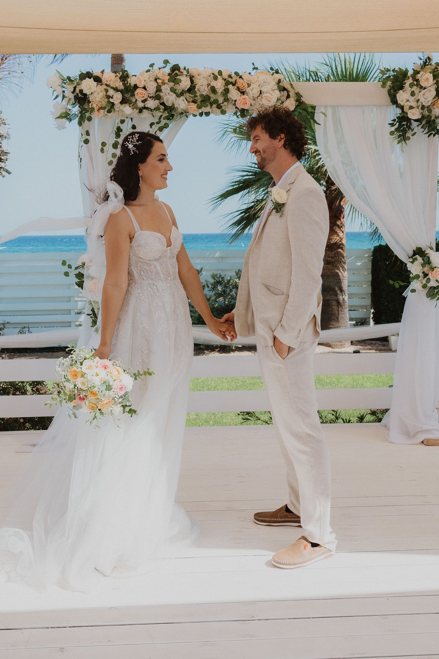 Wedding couple standing under floral arch with ocean and palm trees in background.