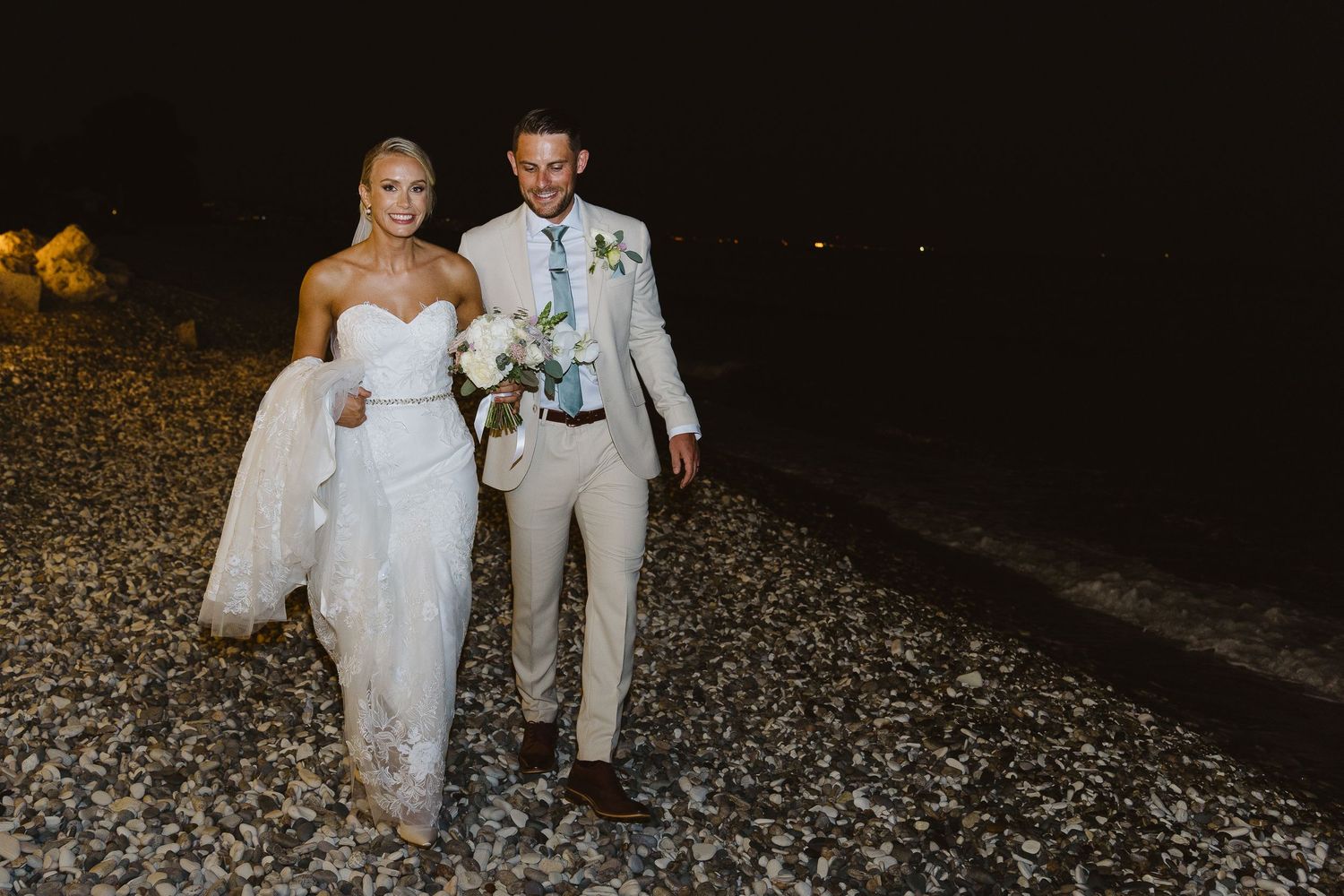A couple walks on a rocky beach at night after their wedding ceremony.