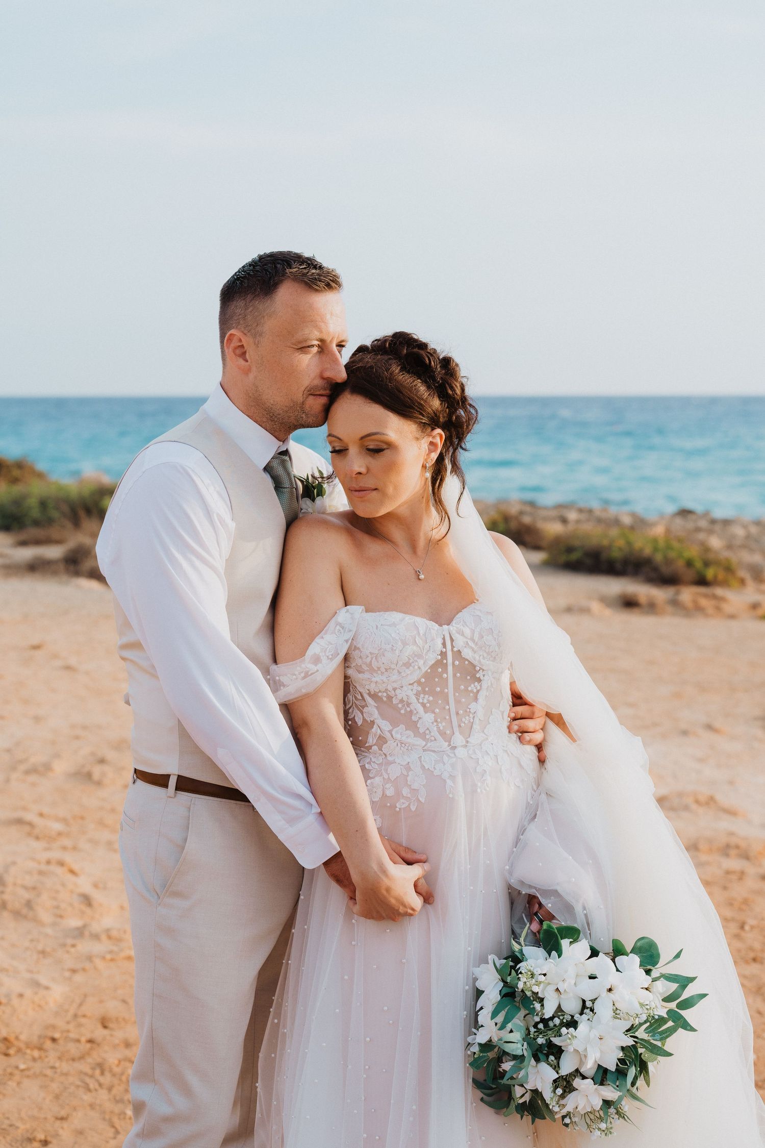 A couple embraces on a beach during their destination wedding, with turquoise ocean waters and a white floral bouquet.