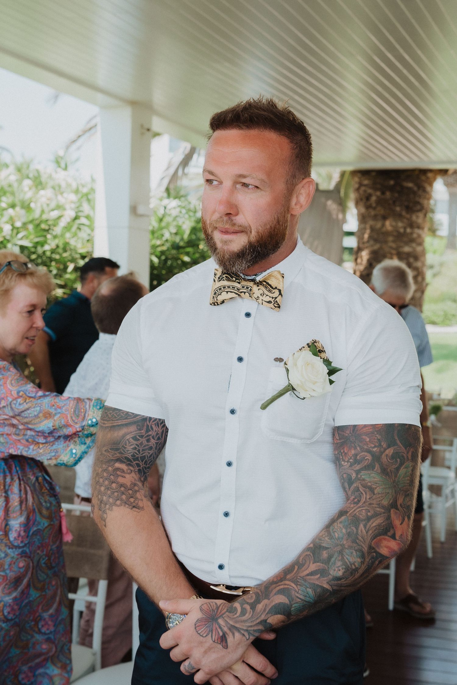 A groom in white shirt with bow tie and tattoos stands during an outdoor wedding ceremony.