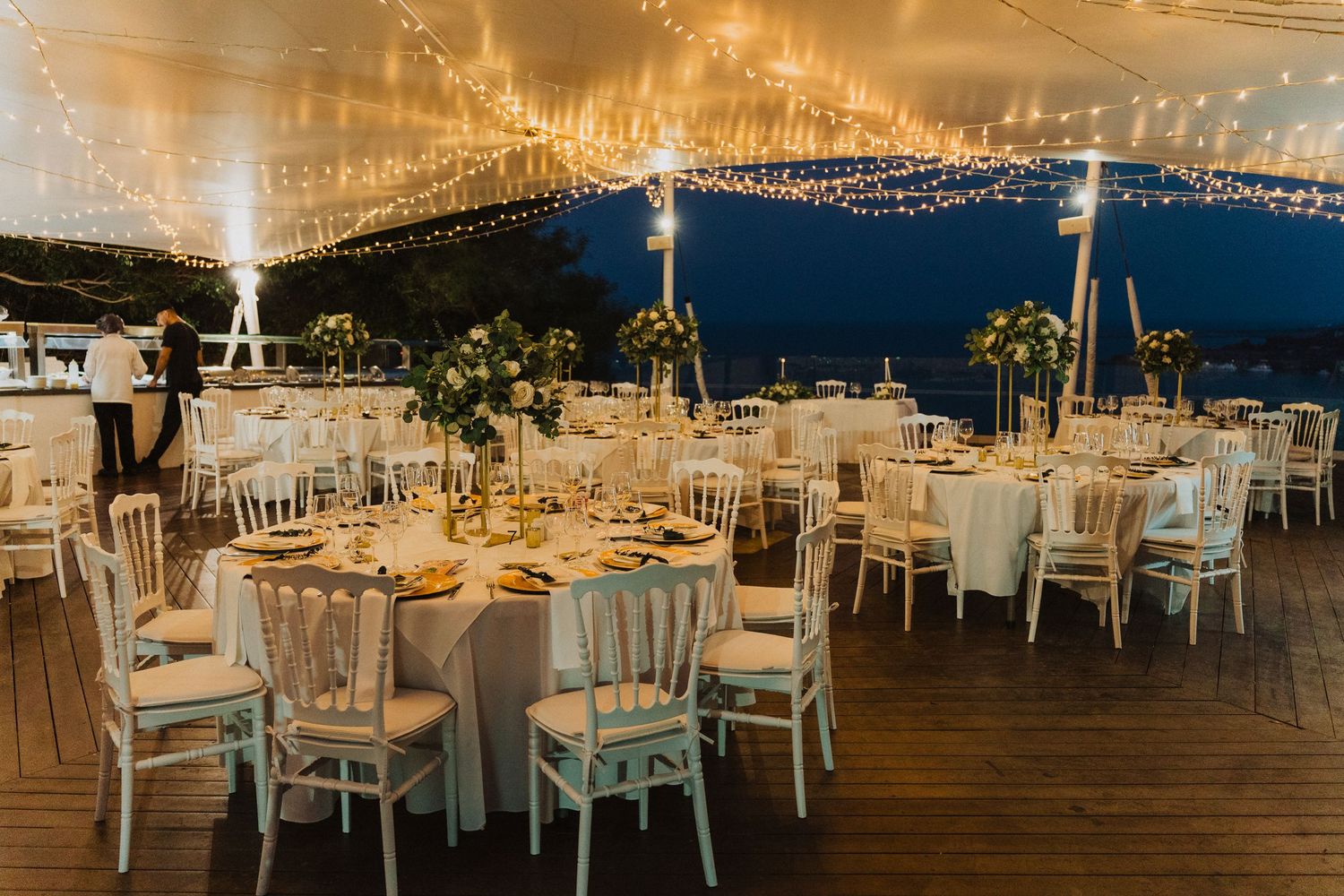 Elegant outdoor wedding reception setup with white tables and chairs under twinkling string lights at dusk.