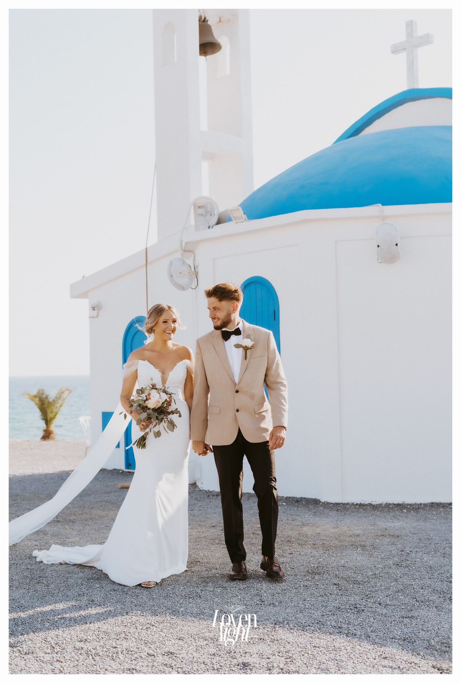 Elegant wedding photo showing couple standing by white Mediterranean church with blue dome.