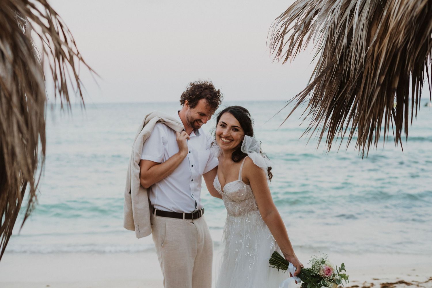 A couple enjoys a romantic moment on a tropical beach under palm fronds during their destination wedding.