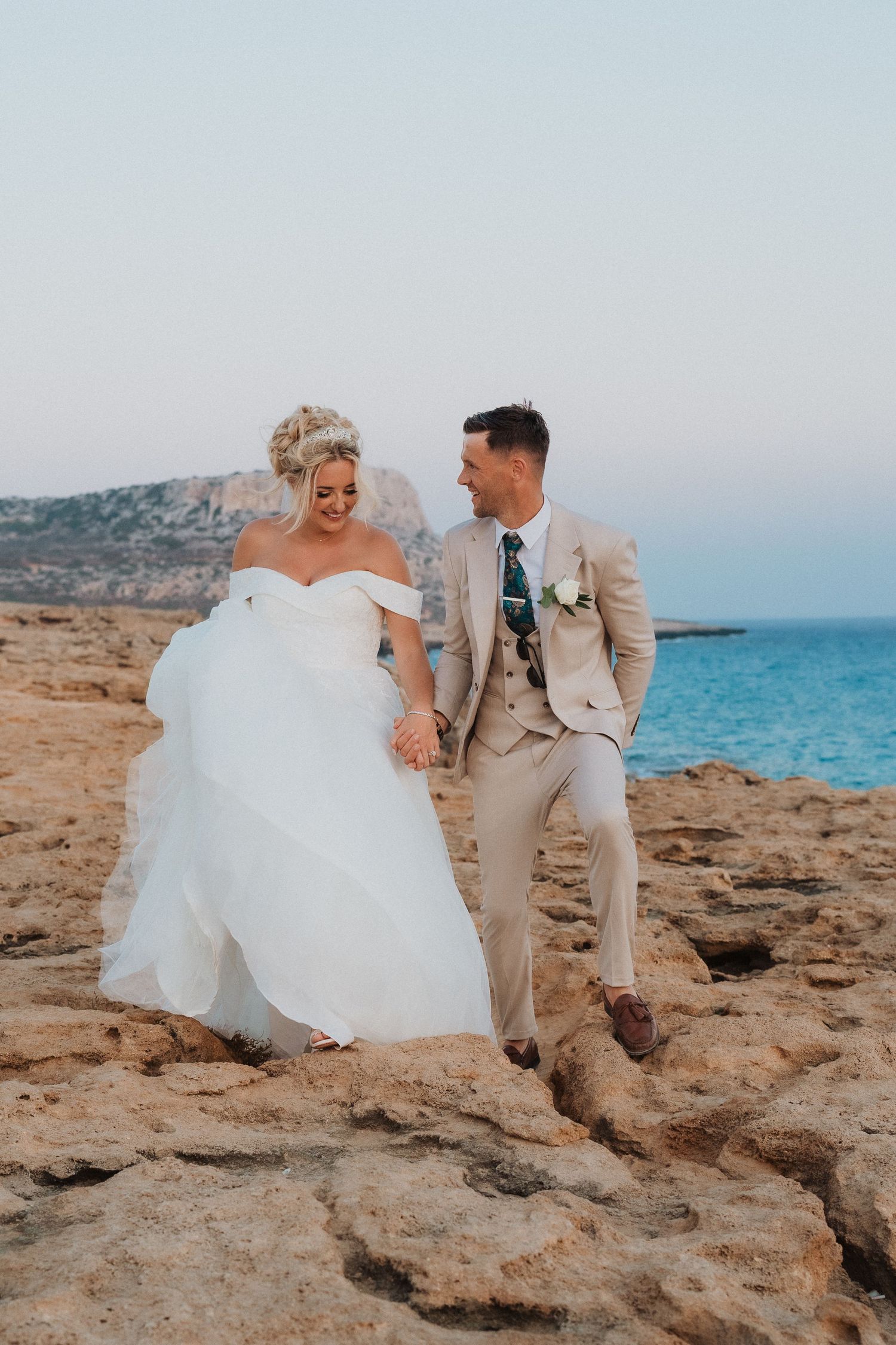Newlyweds share a joyful moment on rocky coastal cliffs overlooking the blue sea.