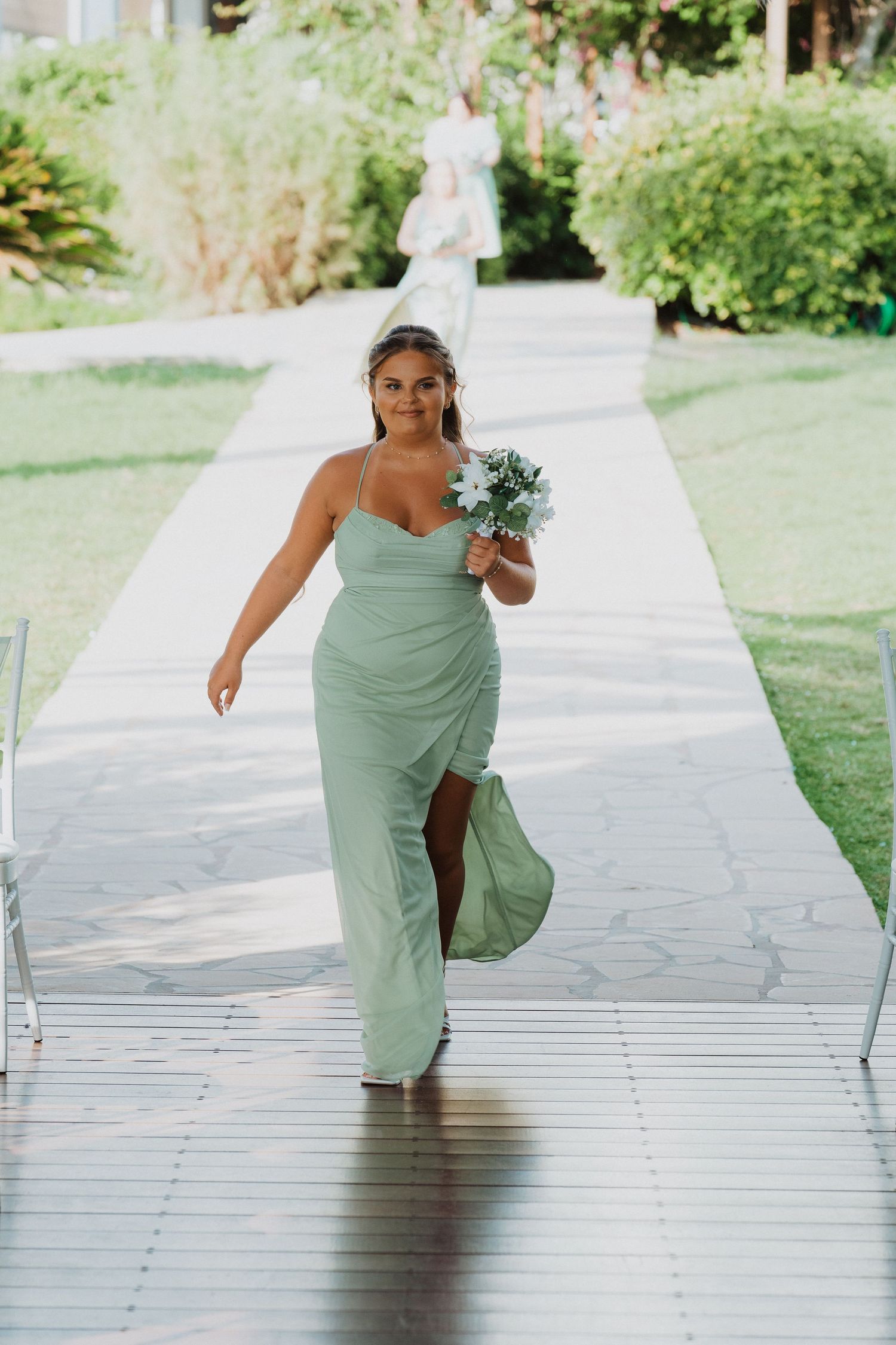 Bridesmaid in sage green slit dress poses on garden walkway while holding floral bouquet.