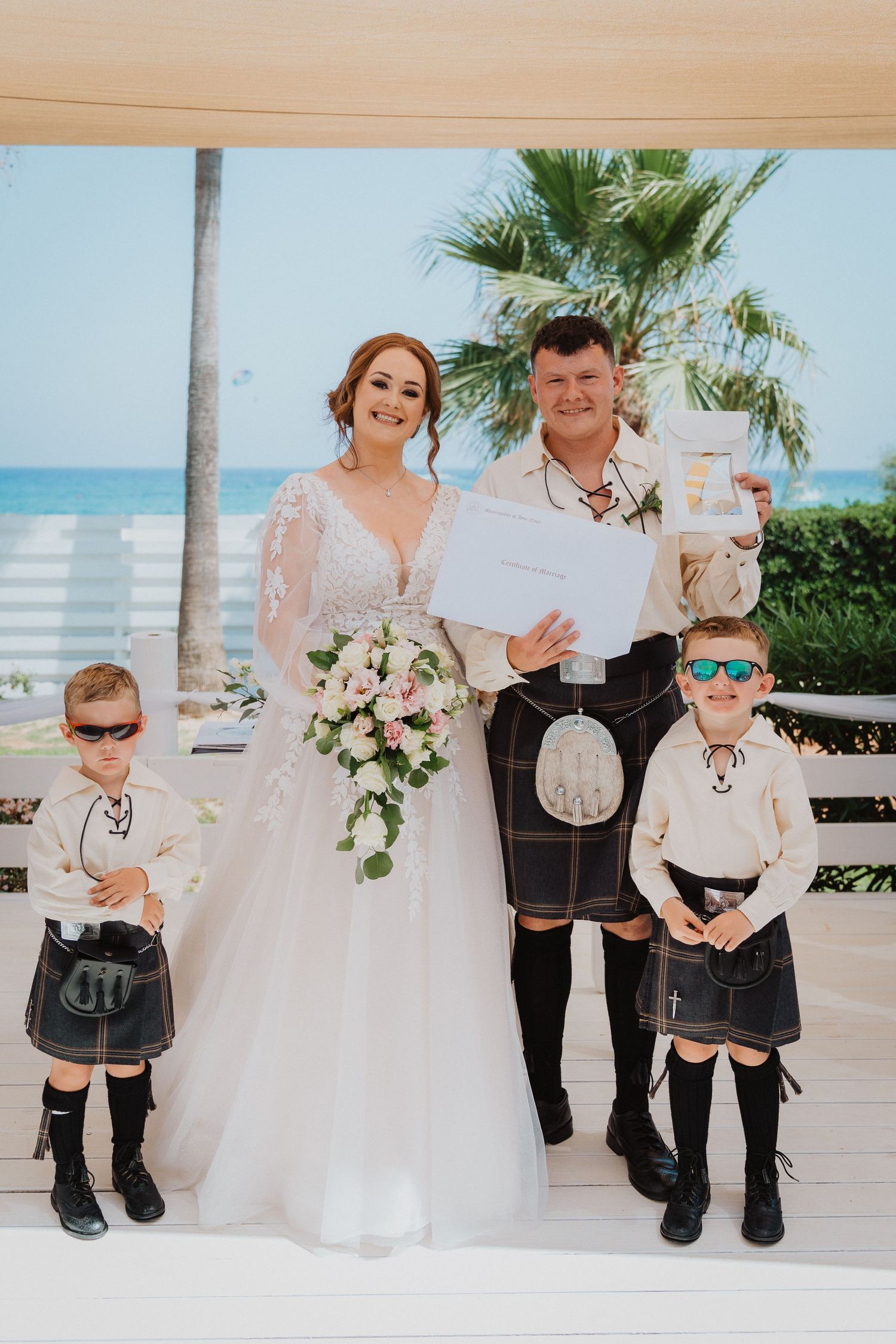 Scottish wedding couple pose with two young children in kilts by the beach with palm trees in background.