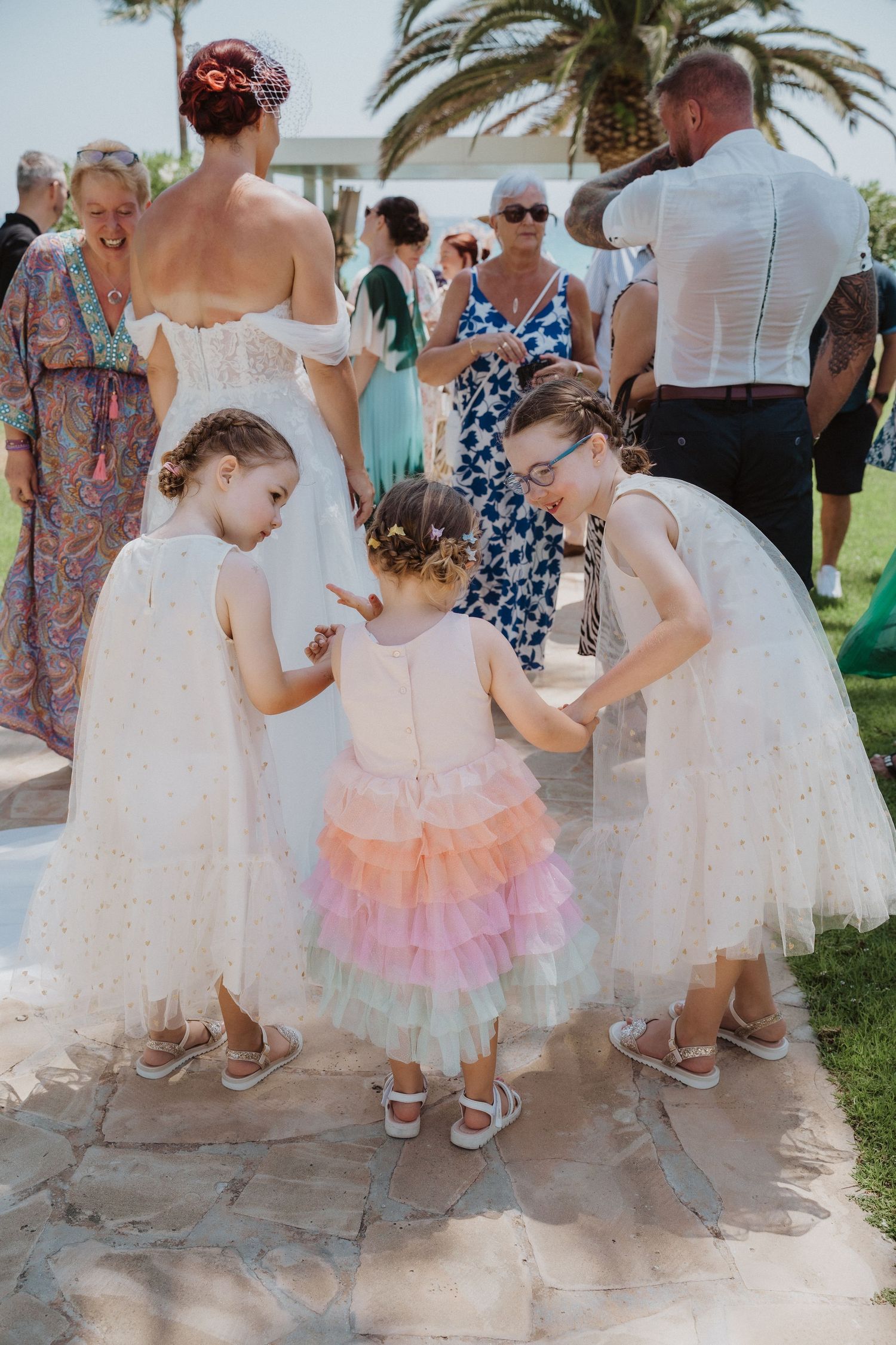 Young flower girls in white dresses interact during outdoor wedding celebration.