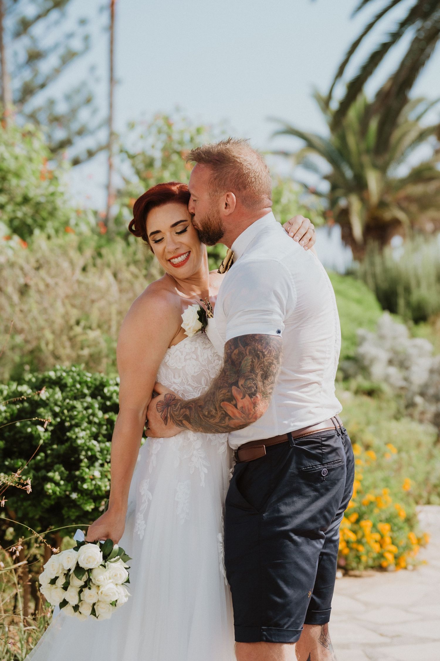 Romantic outdoor wedding scene with couple embracing among palm trees and flowers.