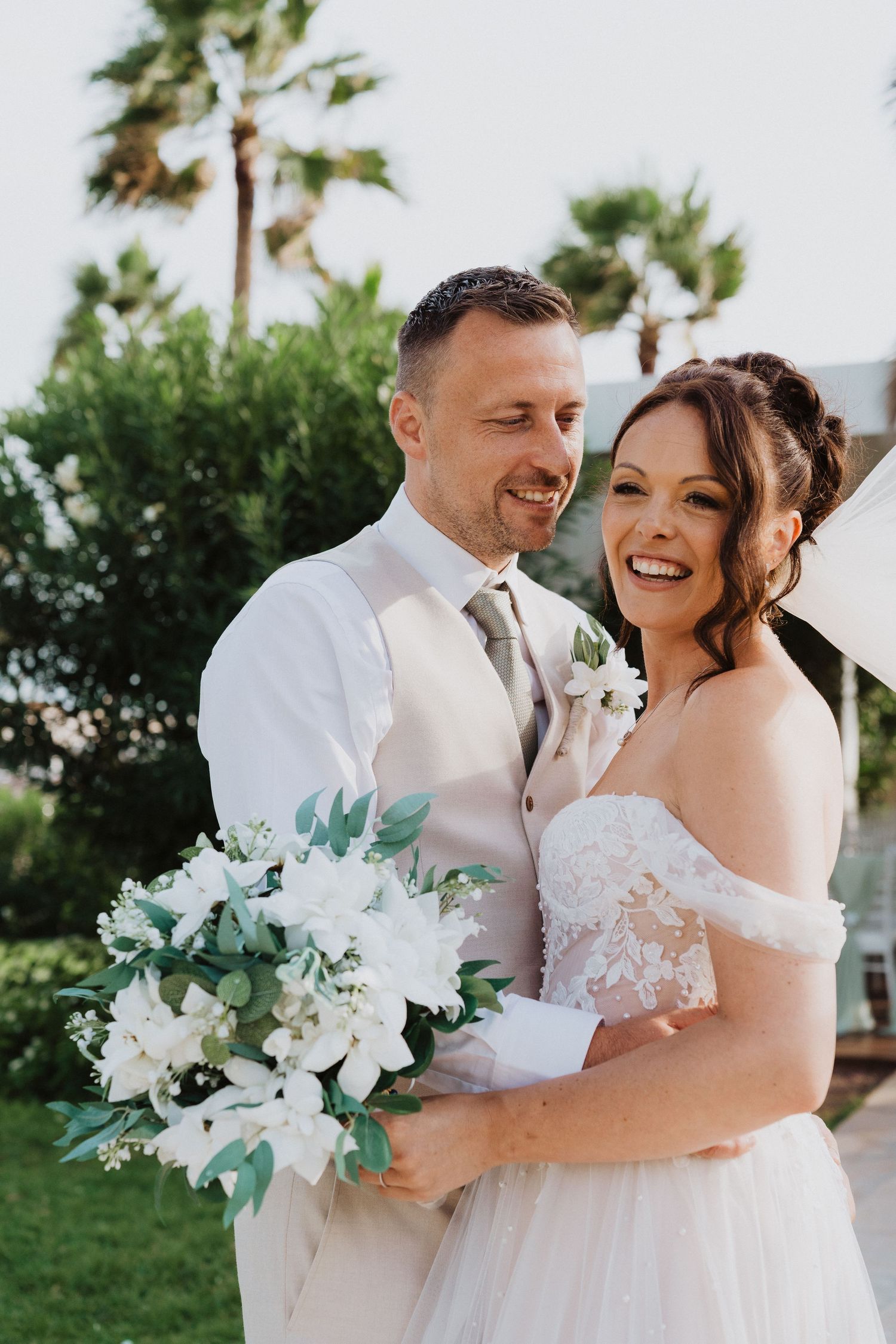 A joyful couple shares a candid moment during their outdoor wedding, with a white floral bouquet and palm trees in the background.
