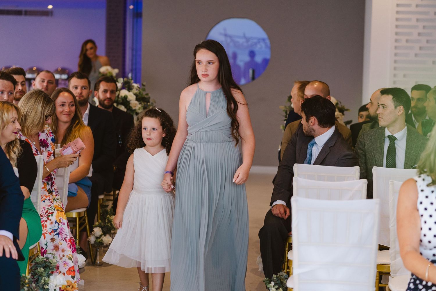 Wedding processional in a modern venue with blue lighting and guests lining the aisle.